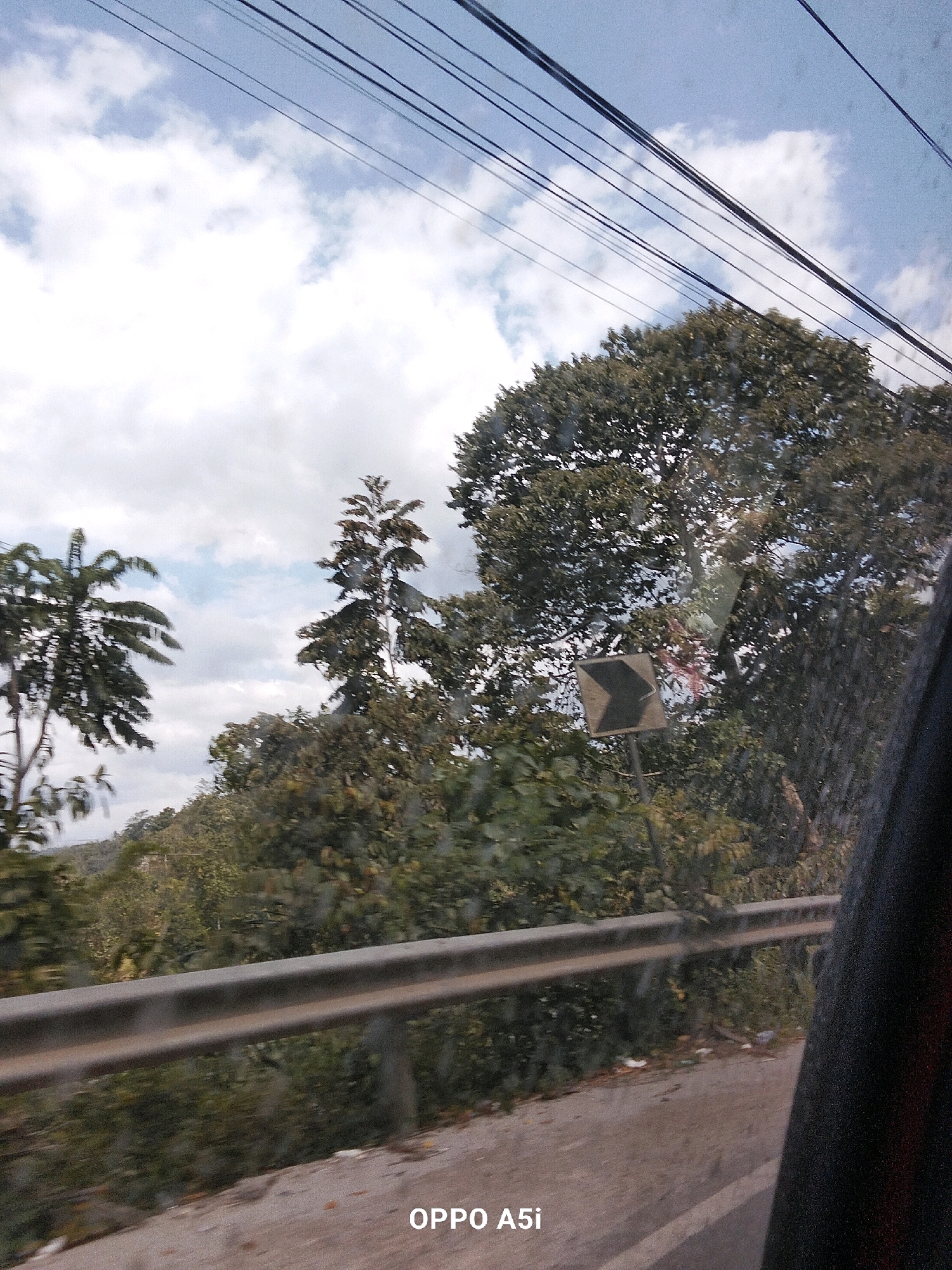 The image shows a roadside view on a cloudy day, likely in Andadowi, Indonesia. The perspective is from a moving vehicle, indicated by the motion blur and the presence of overhead power lines and a sliver of the vehicle's interior on the right. A metal guardrail runs along the side of the road in the foreground. Beyond the guardrail, dense vegetation and trees form a natural backdrop. A diamond-shaped traffic sign with a black chevron pointing to the right is visible on a post amidst the greenery. The sky is partly cloudy, with streaks of blue visible between the clouds. The bottom right corner of the image displays the text "OPPO A5i," likely a watermark from the camera used to capture the photo. No people are visible in the scene.