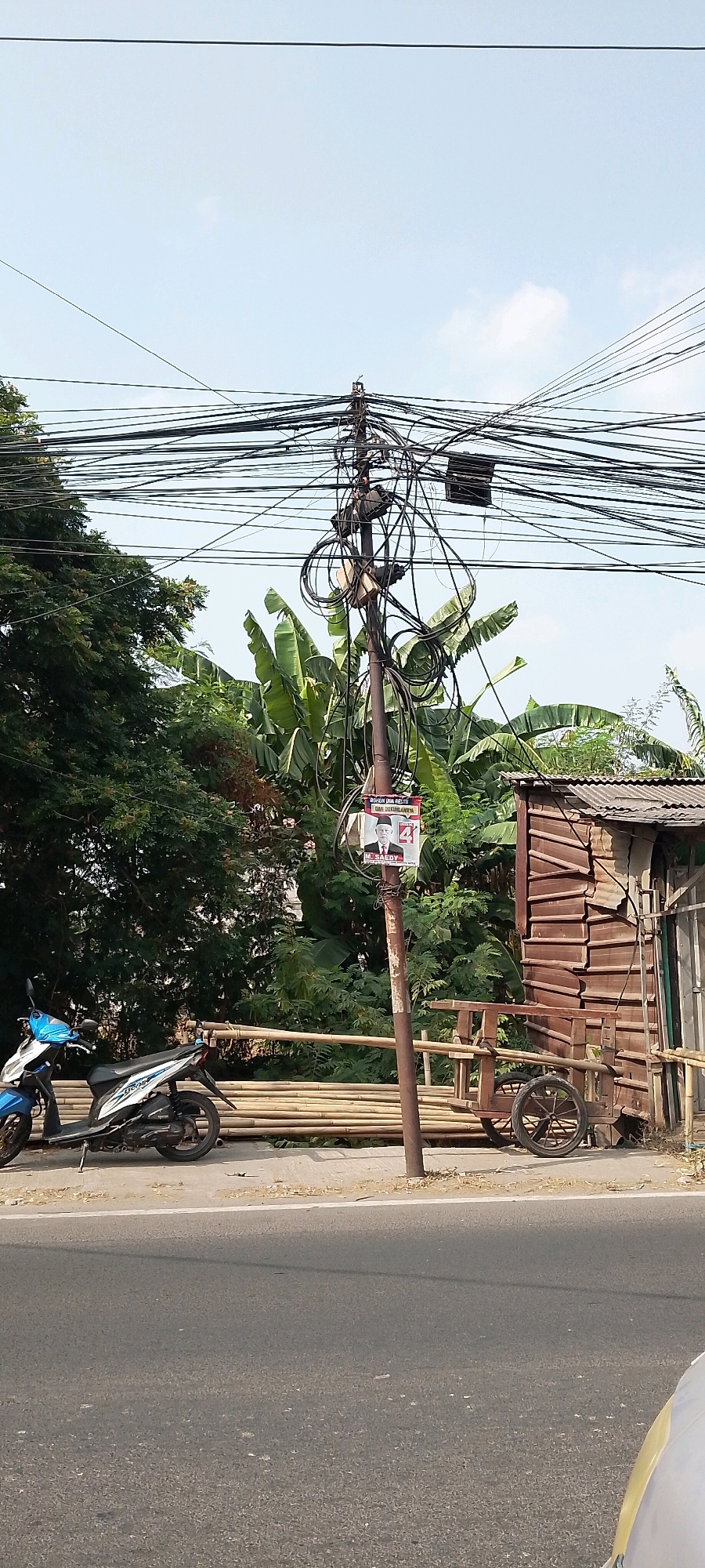 In Karangsatria, Indonesia, a utility pole stands amidst a dense network of overhead electrical wires. Attached to the pole is a poster featuring a man in a suit and a red number "4." To the right of the pole is a rustic wooden structure with a corrugated metal roof, resembling a small shed or stall. A two-wheeled cart with wooden wheels is positioned in front of the structure. In the foreground, a blue and white motorcycle is parked on the side of an asphalt road. The background is filled with lush green trees and foliage, with a clear sky above.