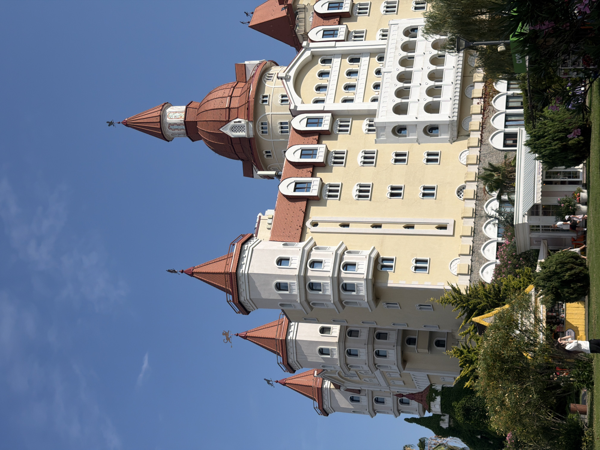 In Sirius, Russia, a striking yellow hotel complex with red-tiled roofs dominates the frame under a clear blue sky. The architecture features multiple turrets, some topped with weather vanes, and a large dome. The building is surrounded by lush greenery, including trees and bushes, and a small yellow kiosk with awnings is visible in the foreground. Several people are present in the scene: a woman in a white shirt stands in the lower-left foreground, taking a photo with her phone, while other individuals are seen walking or standing near the kiosk and in the background. The overall impression is of a sunny day at a resort or hotel.