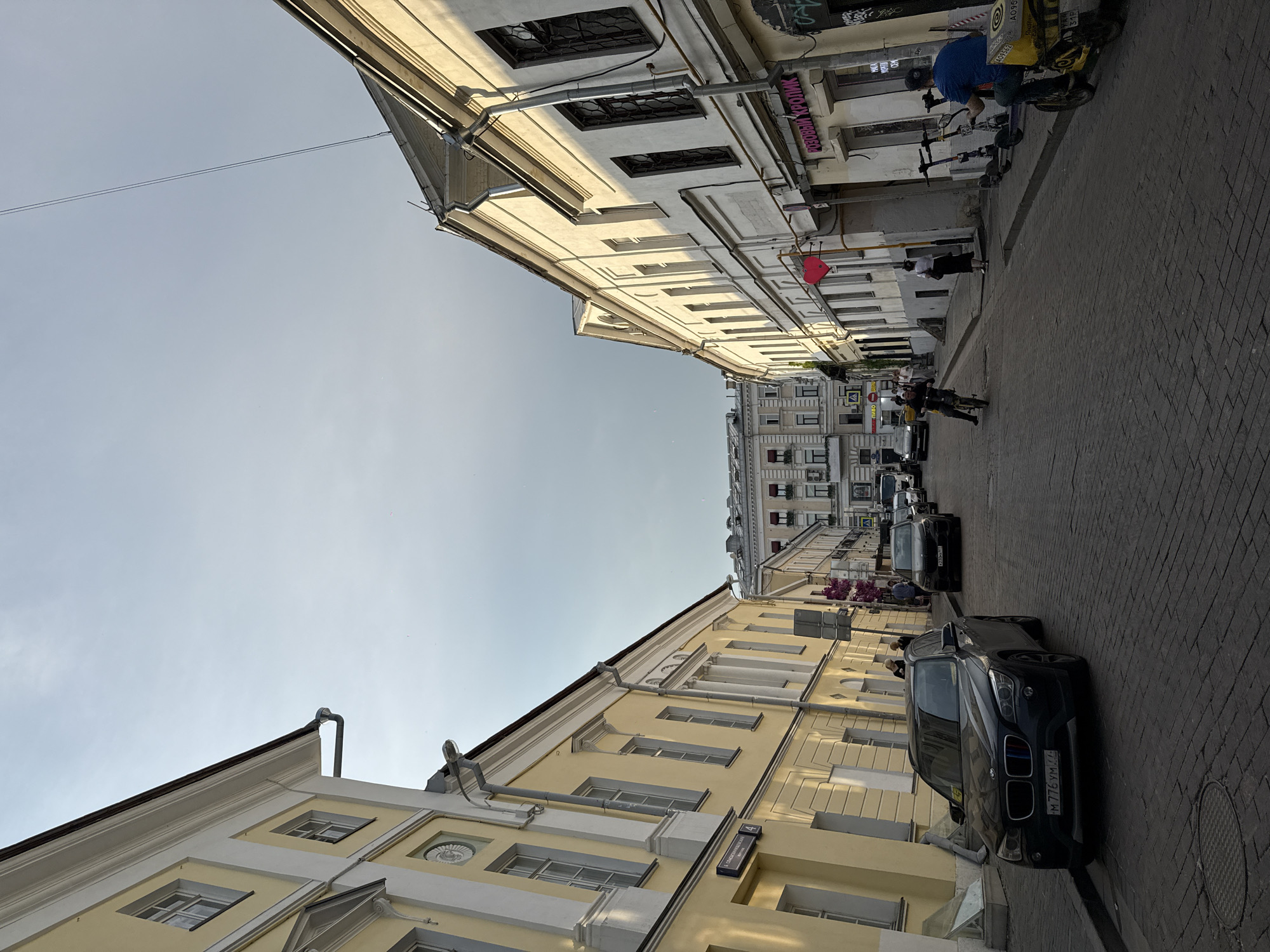 The image captures a street scene in Moscow, Russia. It shows a narrow street lined with buildings on both sides, creating a sense of enclosure. The buildings have a mix of architectural styles, featuring classical elements like columns and decorative moldings. One building is painted in a light yellow color, while the other is white. Cars are parked along the side of the street, including a black BMW SUV. A man on a motorcycle is riding down the street, and there are several scooters parked against one of the buildings. There are also pedestrians walking on the sidewalk. The sky is visible in the space between the buildings, and the overall lighting suggests it is daytime. The street is paved with cobblestones. A sign saying "Садовая-Каретная ул., 4" is seen. The scene conveys a sense of everyday urban life in Moscow.