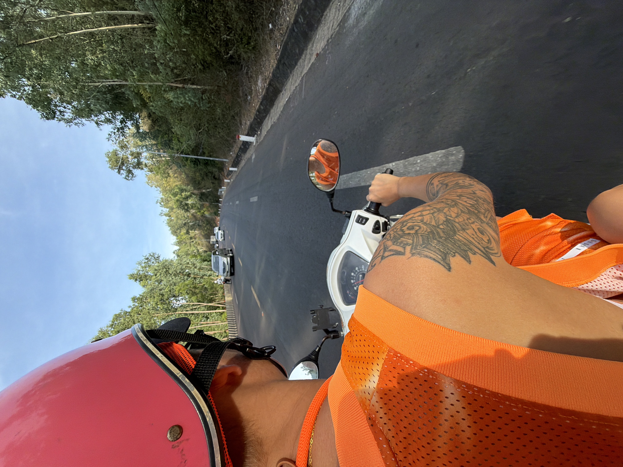 This media file captures a first-person perspective of someone riding a white scooter on a paved road, likely in South Quy Nhon Ward, Vietnam. The rider is wearing a bright red helmet and an orange mesh vest over an orange shirt. A prominent tattoo is visible on the rider's left arm. The scene is set on a road flanked by lush green trees on the left and a clear blue sky overhead. Several vehicles are visible on the road ahead, suggesting moderate traffic. The time of day appears to be daytime, with bright sunlight illuminating the scene. The road has a distinct white line marking the lanes. The overall mood seems relaxed and adventurous, indicative of someone enjoying a ride. No visible text is present in the image.