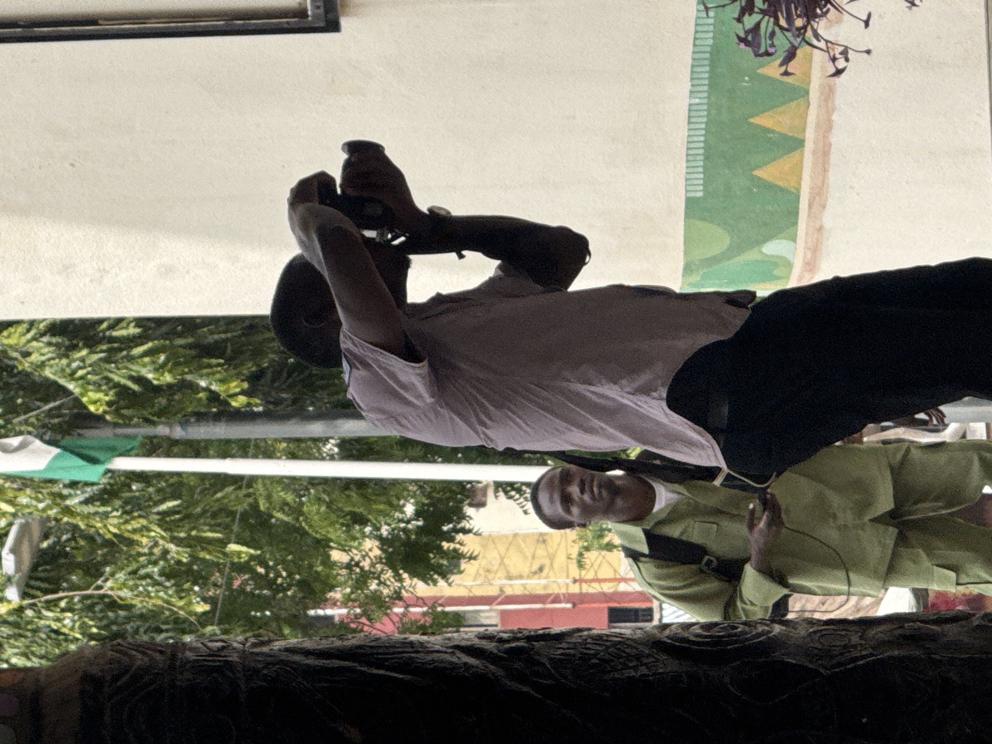The image captures an outdoor scene, likely in Abuja, Nigeria, with a focus on two men and architectural elements. In the foreground, a man in a light pink shirt and dark pants is holding a camera to his face, actively taking a photograph. To his left and slightly behind him, another man wearing a green uniform, possibly a security guard or official, stands looking towards the left. He has a backpack on and appears to be holding a small black object in his hand. Behind the men, a building with a light-colored facade is visible, featuring a decorative band of green and yellow geometric shapes near the bottom. Trees with lush green foliage are present, and a Nigerian flag, predominantly green and white, is seen flying on a pole in the background. The overall impression is that of a public space, possibly near an administrative or cultural building, where photography is taking place.