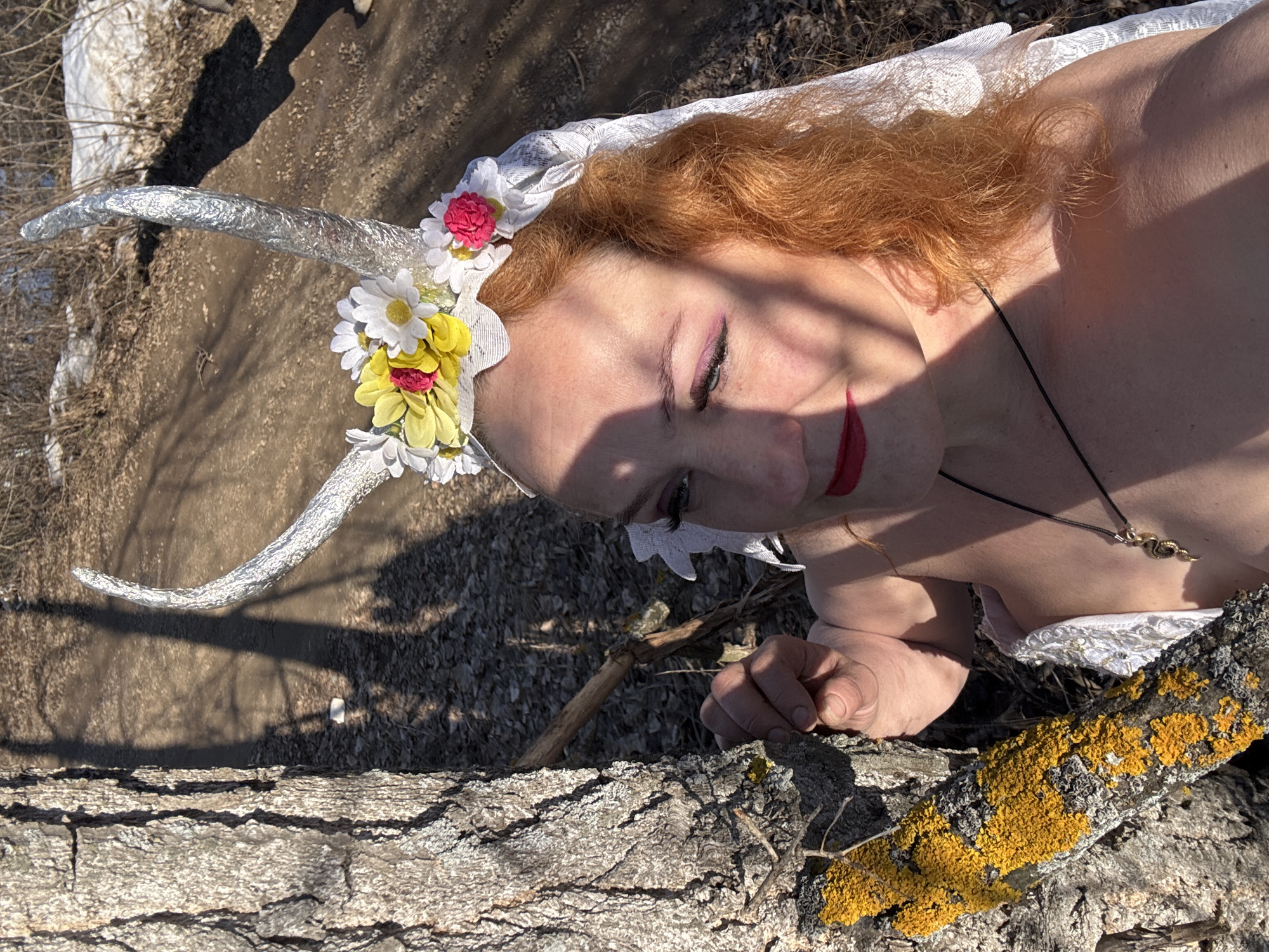A woman lies on a tree trunk covered with lichen in an outdoor setting, possibly in Chaikovsky, Russia. She has red hair, wears a headpiece with floral decorations and silver horns, and has on makeup. She wears a white garment with lace details. The trunk has rough, textured bark and patches of yellow lichen. In the background, there is a path with loose dirt and some visible snow or ice patches. Bare tree branches and shadows cast by the sun are present.