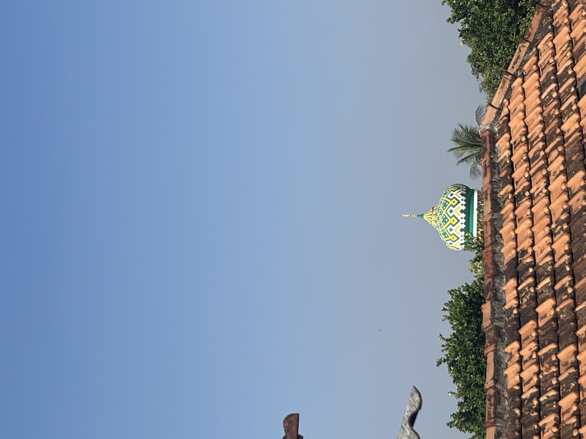 A clear blue sky dominates the upper portion of the image. In the lower half, a tiled roof made of reddish-brown terracotta tiles fills the foreground. Behind the roof, lush green trees are visible. Nestled among the trees and slightly above them is a dome of a mosque, characterized by intricate green and yellow patterned decorations and a golden spire. To the left of the dome, the fronds of a palm tree can be seen. The overall scene suggests a bright, sunny day. The location is Glagah Kulon, Indonesia.