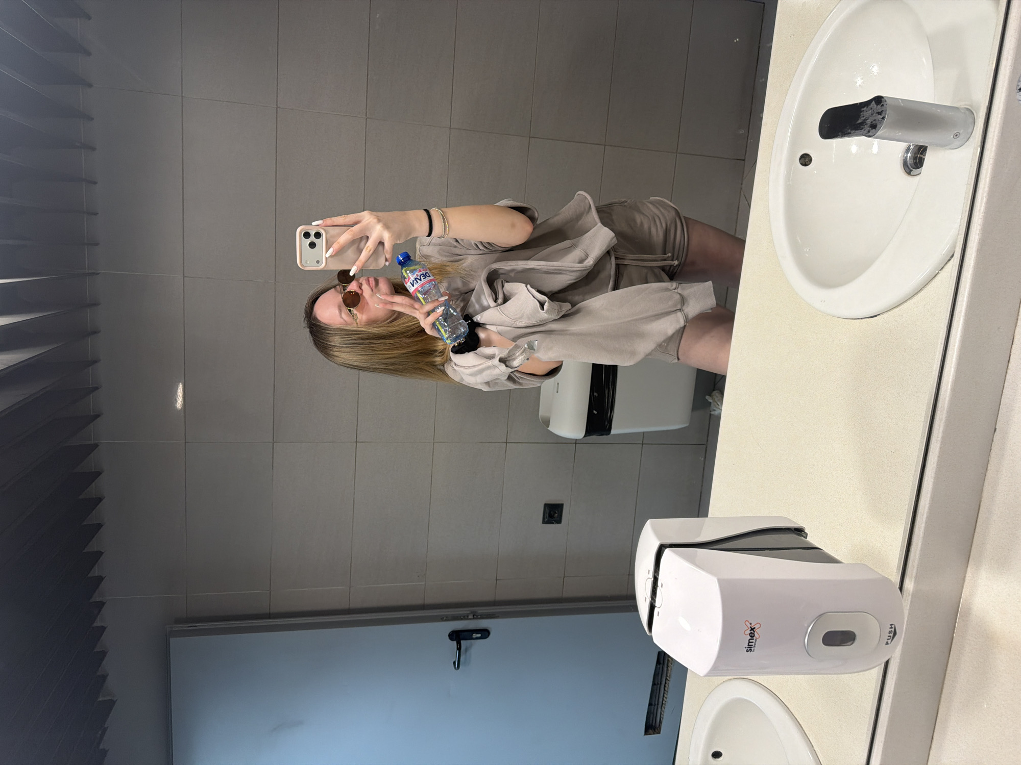 A woman in a tan, two-piece outfit and sunglasses is taking a selfie in a bathroom. She is holding a water bottle in her left hand and making a peace sign. She has blonde hair. The bathroom has grey tiled walls and a white sink. A Simex-branded soap dispenser is located on the counter. A door with a black latch is in the background. The location is Blagoevgrad, Bulgaria.