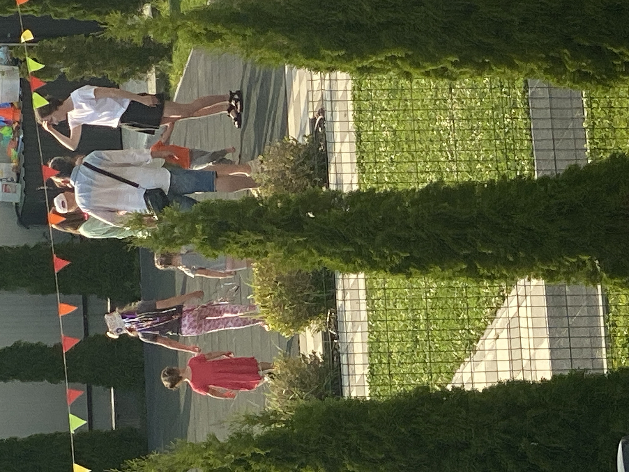 In Novi Petrivtsi, Ukraine, a group of people are gathered outdoors, possibly for an event or celebration, indicated by the colorful triangular flags strung up. The scene is bathed in the warm light of what appears to be late afternoon or early evening. Several individuals are visible, including a person in a white t-shirt and dark shorts, another in a blue and white shirt with a black crossbody bag, and a person in a red dress. They are standing on a paved area adjacent to a well-maintained green space, possibly a lawn or artificial turf, enclosed by a wire mesh fence. Cypress trees or similar greenery line the perimeter of the scene, partially obscuring the view. The overall atmosphere seems casual and relaxed, with people interacting and observing their surroundings. The ground is paved with rectangular tiles. The specific event is unclear, but the presence of decorations and the gathering of people suggest a social occasion.