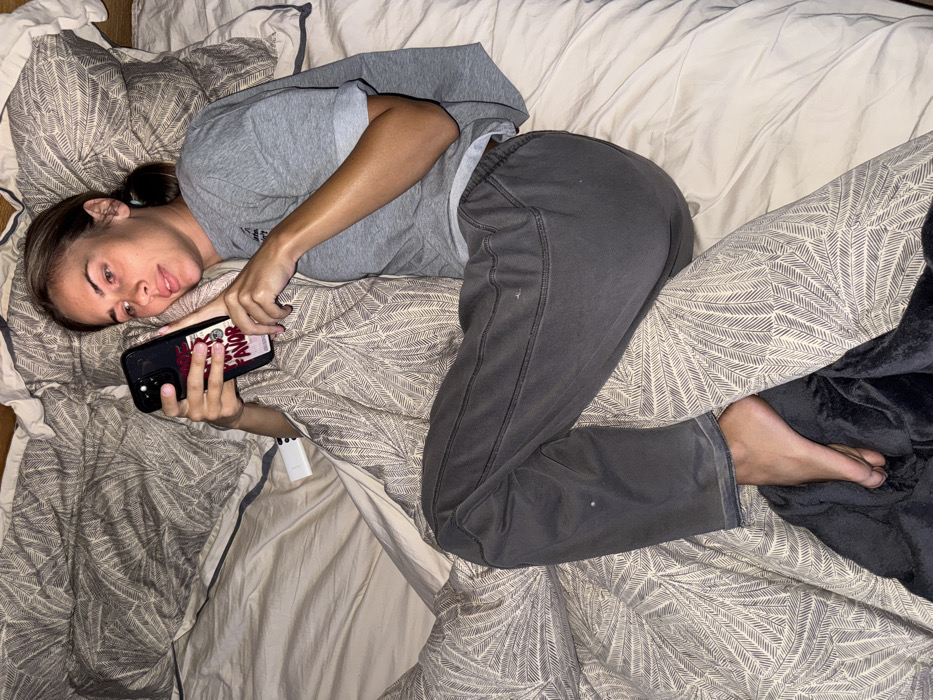 A young woman lies on her back in a bed with patterned bedding. She wears a gray t-shirt and gray sweatpants, and her hair is pulled back. She holds a phone with a dark case and prominent red nails. A white object is visible near her hand. A corner of a dark blanket is at the edge of the frame near her foot. The room is softly lit.