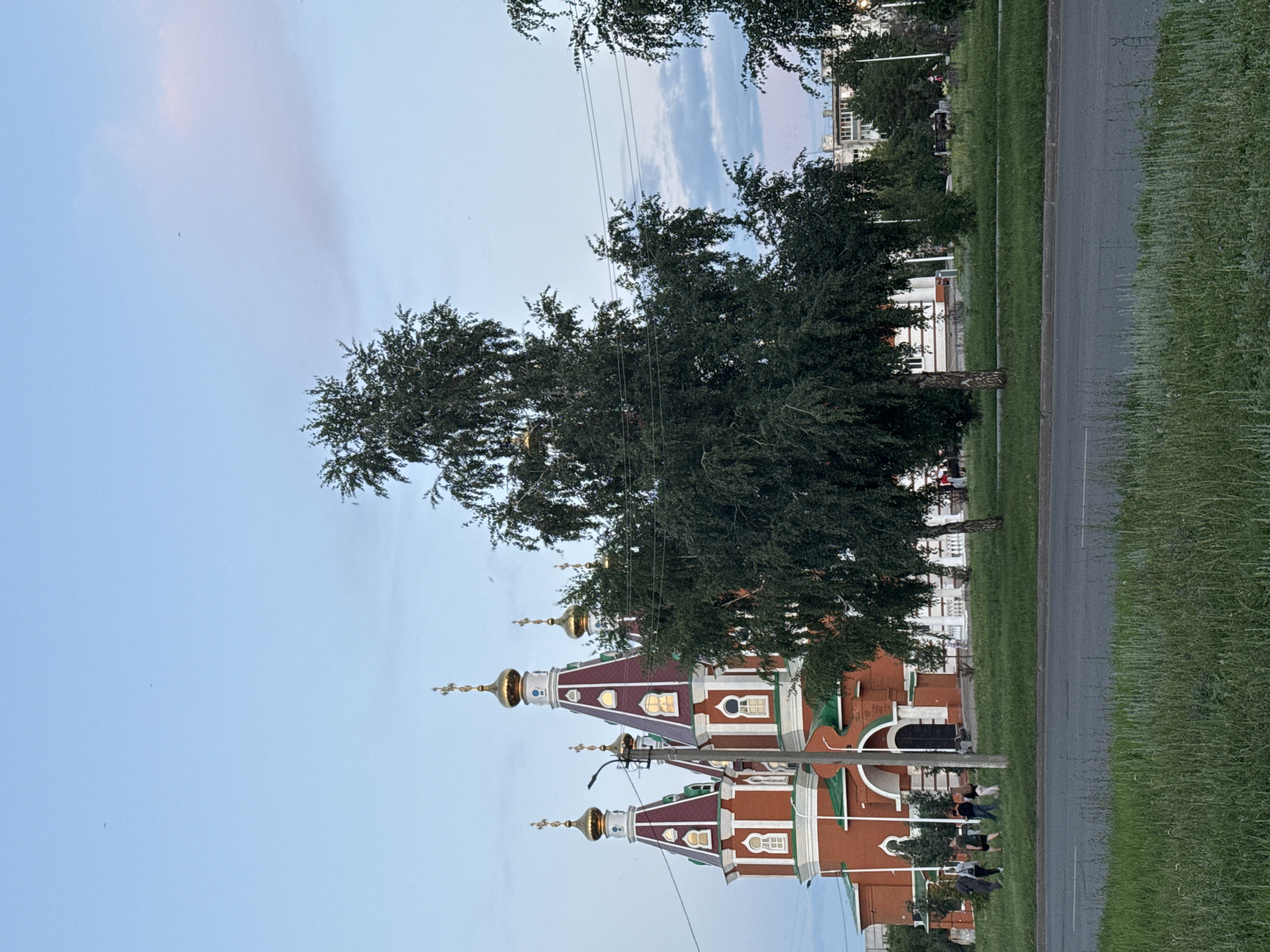 In Otradny, Russia, a red brick church with multiple gold-domed towers is visible. The church features red roofs with white trim and decorative windows. In the foreground, a large deciduous tree with dark green foliage partially obscures the view of the church. Beyond the church, there are other trees, a building, and telephone wires. Several people are walking on the grass area in front of the church. A paved road with grass on the verge runs across the bottom of the image. The sky is a pale blue with hints of pink in the distance, suggesting it is late afternoon or early evening.