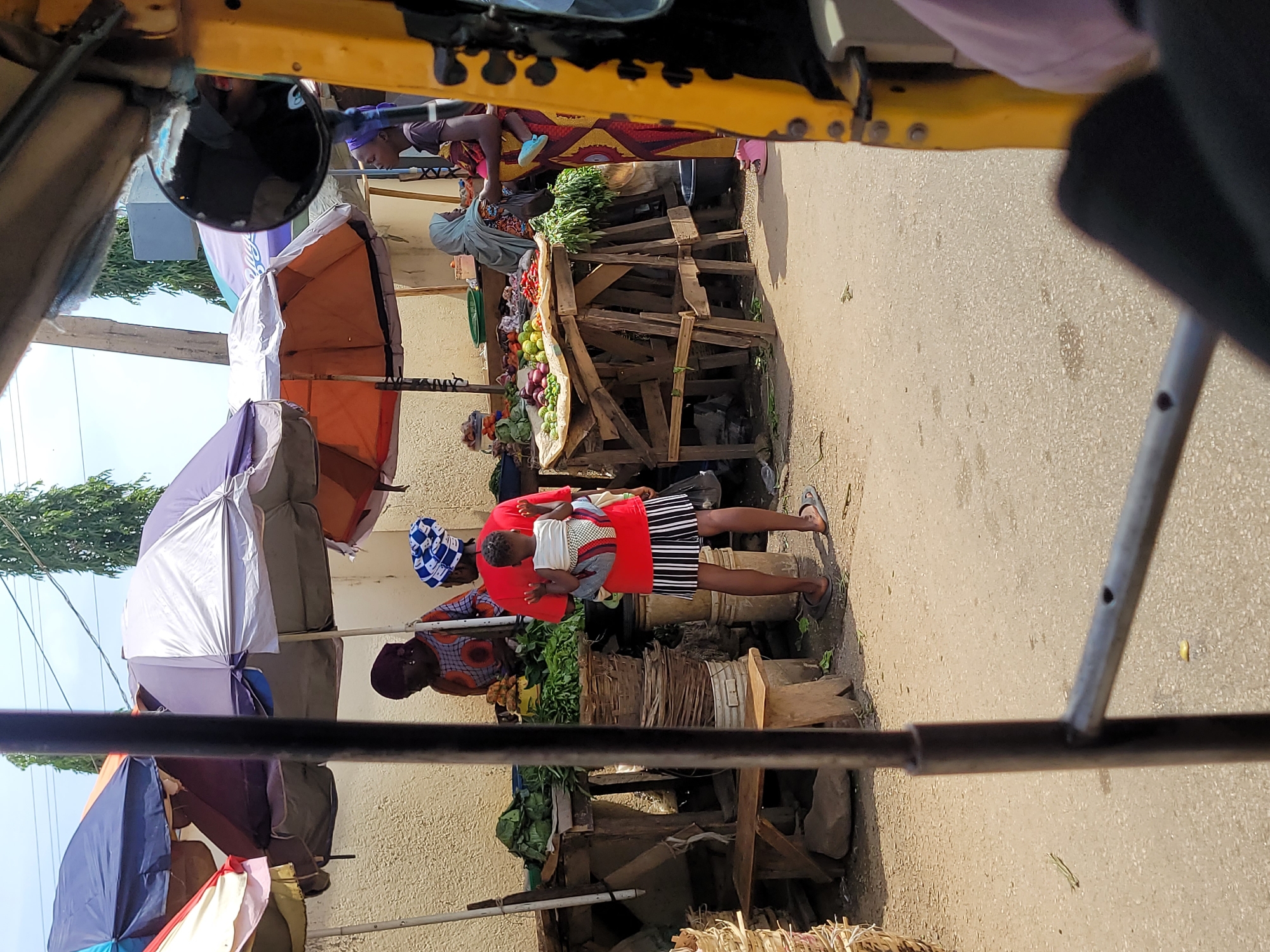 The image captures a vibrant street market scene in Jos, Nigeria, viewed from the perspective of someone inside a vehicle, likely a tricycle, whose yellow frame is visible at the top and bottom edges of the photograph.
In the center of the frame, a woman stands prominently, wearing a bright red top and a black and white striped skirt. She carries a baby on her back, securely wrapped in a patterned fabric, a common practice in Nigeria. The woman is positioned amidst several makeshift market stalls constructed from rough-hewn wood. These stalls are laden with an array of fresh produce, including abundant leafy green vegetables, onions, and other fruits and vegetables, indicating a bustling food market.
Behind the central figure and further into the market, several other women are visible, appearing to be vendors or shoppers. One woman wearing a light blue headwrap is attending to a stall filled with produce. To provide shade from the bright daylight, multiple large, colorful umbrellas in shades of orange, white, purple, and blue are erected over the market stalls. A side-view mirror from the vehicle is also partially visible on the left side of the image. The ground is a paved surface, suggesting a street or open area dedicated to the market. The bright, clear lighting indicates it is daytime. No discernible text is visible in the image.