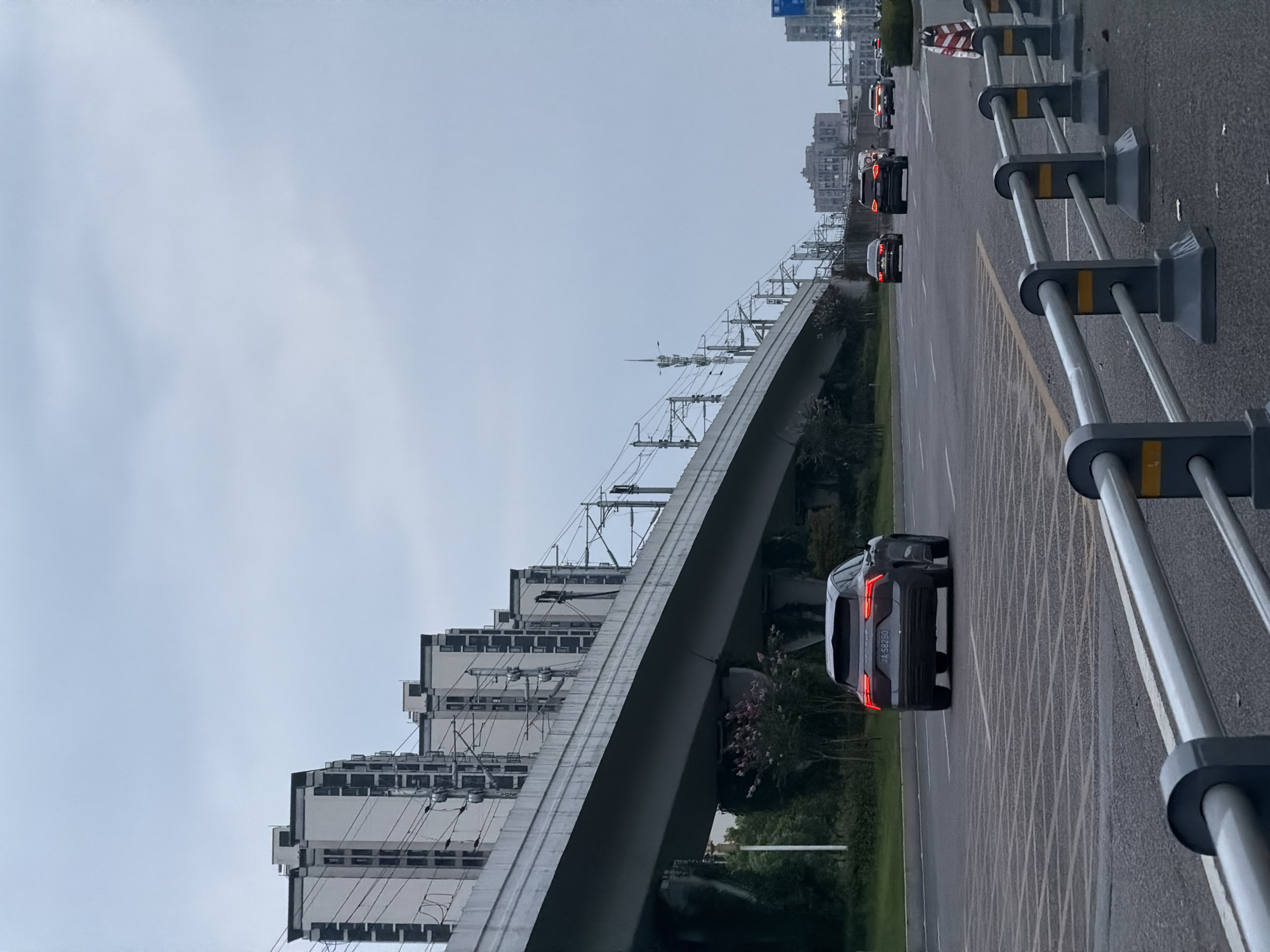 The image depicts a street scene in the city of Jiangjin, China. In the foreground, a gray SUV with license plate "A 58260" is driving on an asphalt road. The SUV has its brake lights illuminated. The road is marked with white lines. Beyond the SUV, other vehicles are visible, including a black sedan and several SUVs. In the background, an elevated structure, likely a bridge or overpass for a railway, spans across the scene. The structure is supported by concrete pillars and carries overhead wires for what appears to be an electric railway. On the left side of the image, several tall, modern apartment buildings with dark-framed windows are situated behind the elevated structure. The sky is overcast and gray, suggesting a cloudy day. The overall environment appears to be an urban or suburban setting with significant infrastructure for transportation.