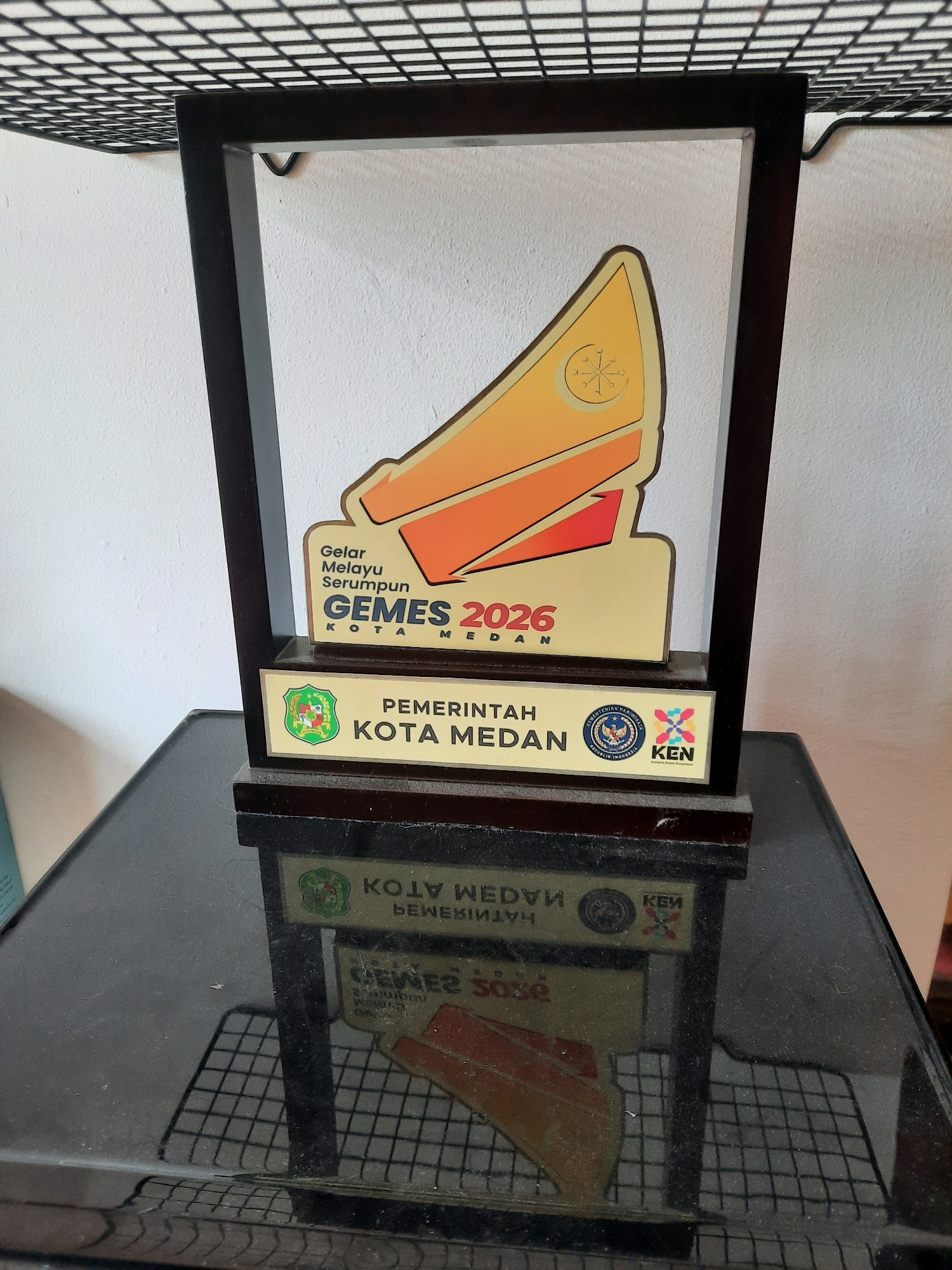 A trophy is displayed on a reflective black surface. The trophy has a dark wooden frame and a light-colored plaque with text and graphics. The text on the plaque reads "GELAR MELAYU SERUMPUN GEMES 2026 KOTA MEDAN" and "PEMERINTAH KOTA MEDAN." There are two circular logos on the right side of the plaque, one with an eagle and text that appears to be related to the Republic of Indonesia, and another with colorful geometric shapes and the letters "KEN". The trophy is positioned in front of a white wall. The reflective surface below the trophy shows a reflection of the trophy and a grid pattern, possibly from a shelf or rack above. The image is taken from a slightly elevated perspective. The scene does not provide any specific information about a location in the city of East Jakarta, Indonesia.