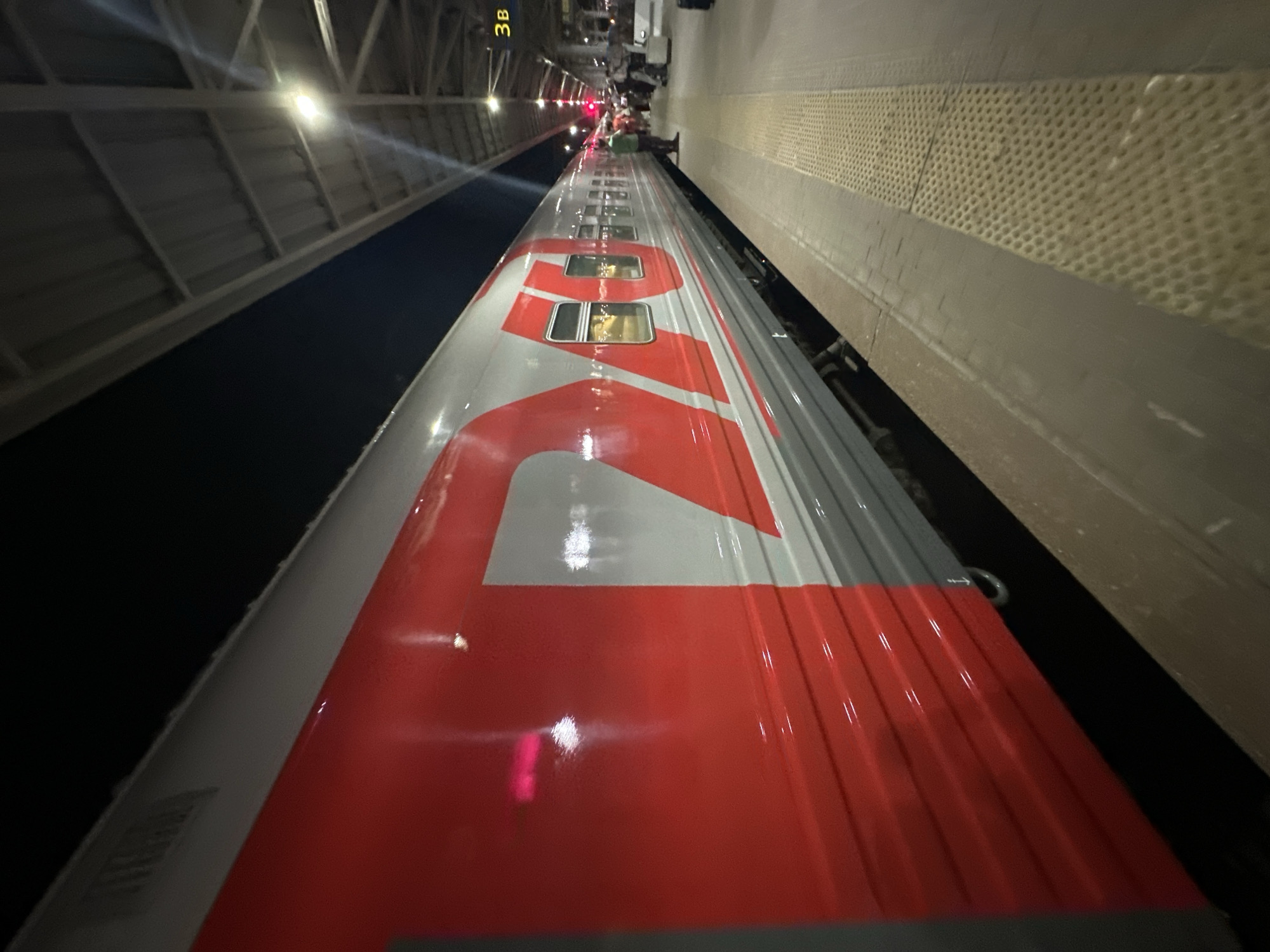 The image shows the roof of a train, likely a Russian Railways (RZD) train based on the distinct red and gray color scheme. The train is stopped at a station platform, evidenced by the textured platform edge on the right side of the frame. A covered walkway or roof structure is visible on the left. In the distance, a sign displaying "3B" is visible above the train. Given the context, it is highly likely that this image was taken at the Moskovsky railway station in Nizhny Novgorod, Russia, as it is a major hub for RZD trains. No people are clearly visible interacting with the train, although a blurry figure can be seen in a window. The lighting suggests it is nighttime.