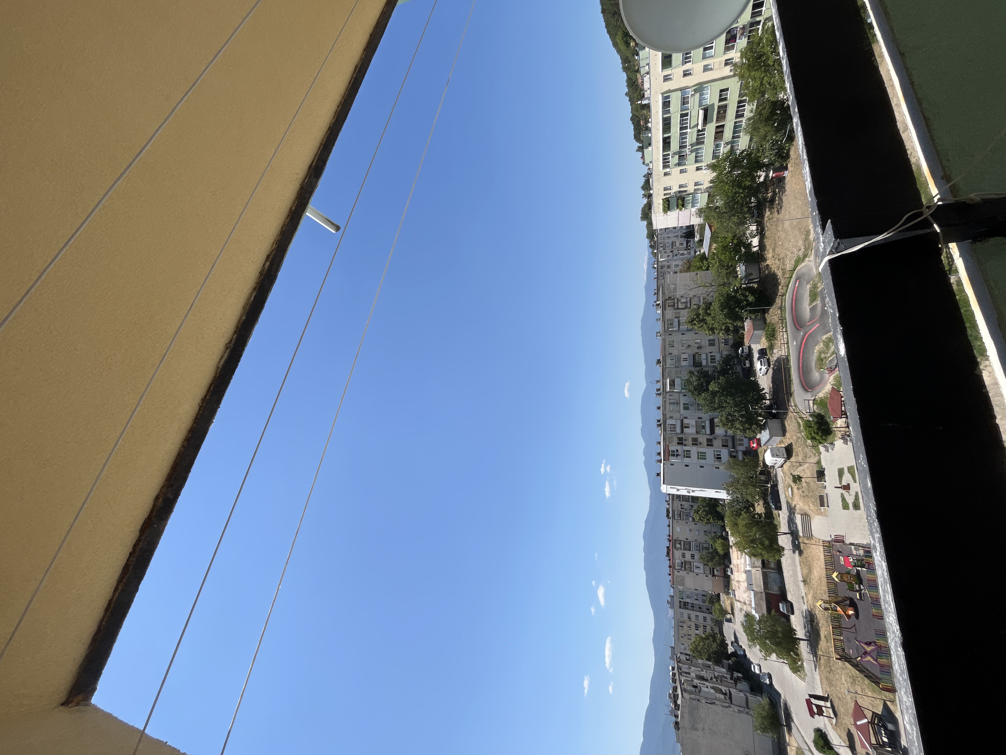 The image captures a view from a balcony overlooking a residential area in Sopot, Bulgaria. In the foreground, a black railing is visible, with some white wires attached. Above this, a beige awning partially obscures the bright blue sky, which is largely clear, with a few small white clouds in the distance.
The middle ground features a collection of multi-story apartment buildings, characteristic of Soviet-era architecture, interspersed with green trees and some smaller structures. A playground with colorful equipment is present, alongside a paved area with benches and a small gazebo. A skate park with red and grey curves is also visible. Cars are parked on the street and in parking areas.
The background is dominated by a range of hazy, blue-green mountains. The overall scene depicts a sunny day in a city with a mix of urban development and natural landscape. There are no people clearly identifiable in the image.