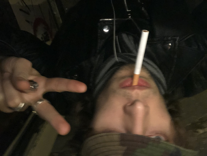 A person with a cigarette jutting from their lips is captured in a tilted, close-up shot. The individual wears a dark jacket and a camo-patterned garment around their neck. Visible rings adorn the fingers of a hand making a "peace" sign. The overall dark and grainy aesthetic contributes to an edgy, candid feel.