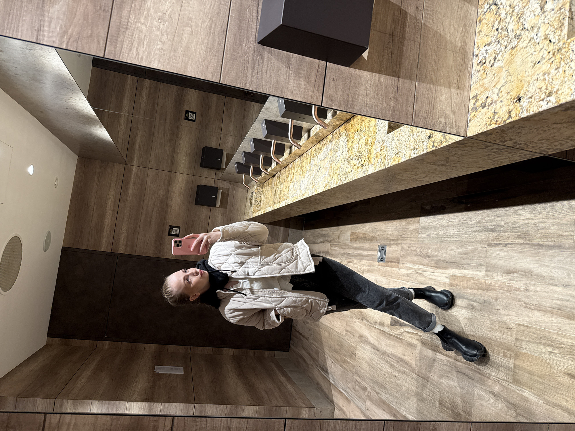 A woman takes a mirror selfie in a bathroom. The woman is wearing a beige quilted jacket, a white top, black pants, and black boots. She is holding a pink phone in her right hand. The bathroom has wood paneling on the walls and floor. There is a mirror above a sink with a granite countertop. There are several soap dispensers on the wall next to the sink. A black box is mounted above the soap dispensers. A round vent is visible in the top left corner of the mirror.
