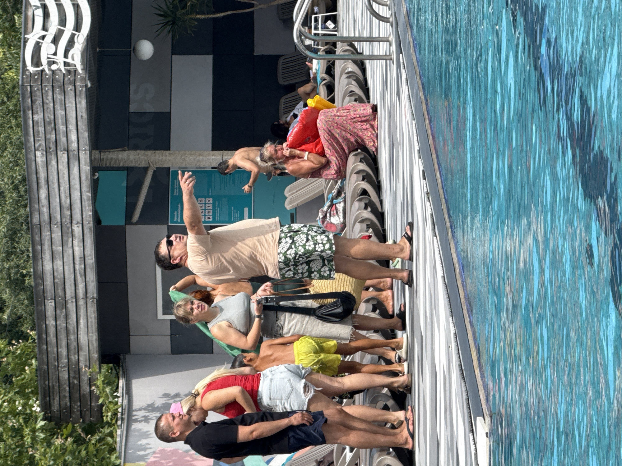The image depicts a poolside scene with several individuals. In the foreground, a bright blue swimming pool occupies the lower half of the frame. The poolside area features a patterned walkway and lounge chairs. Several people are standing and interacting near the walkway. One man, wearing sunglasses and a light-colored t-shirt and patterned shorts, is pointing to the right. A woman in a grey tank top and a light-colored skirt is holding a black purse. A couple stands to the left, a man in a black t-shirt and dark shorts, and a woman in a red tank top and ripped denim shorts. A young boy in yellow shorts stands between them. Further back, a woman in a pink patterned dress is sitting on a lounge chair and appears to be inflating a red object. Another shirtless man is visible in the background. The setting appears to be an outdoor leisure area, possibly a resort or hotel, with some foliage and architectural elements suggesting a built environment. The location is identified as Массандровский поселковый совет, Ukraine.