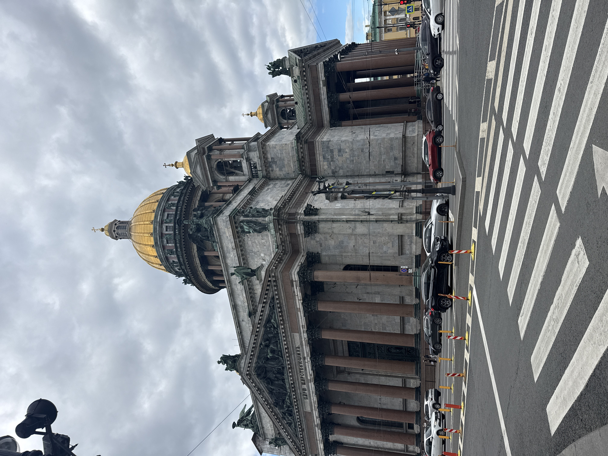The image shows St. Isaac's Cathedral in Saint Petersburg, Russia, from a low-angle perspective. The cathedral's golden dome is prominent, along with its gray stone facade and Corinthian columns. A crosswalk is in the foreground of the cathedral, with parked cars. The sky is cloudy. The time of the image is during the daytime.