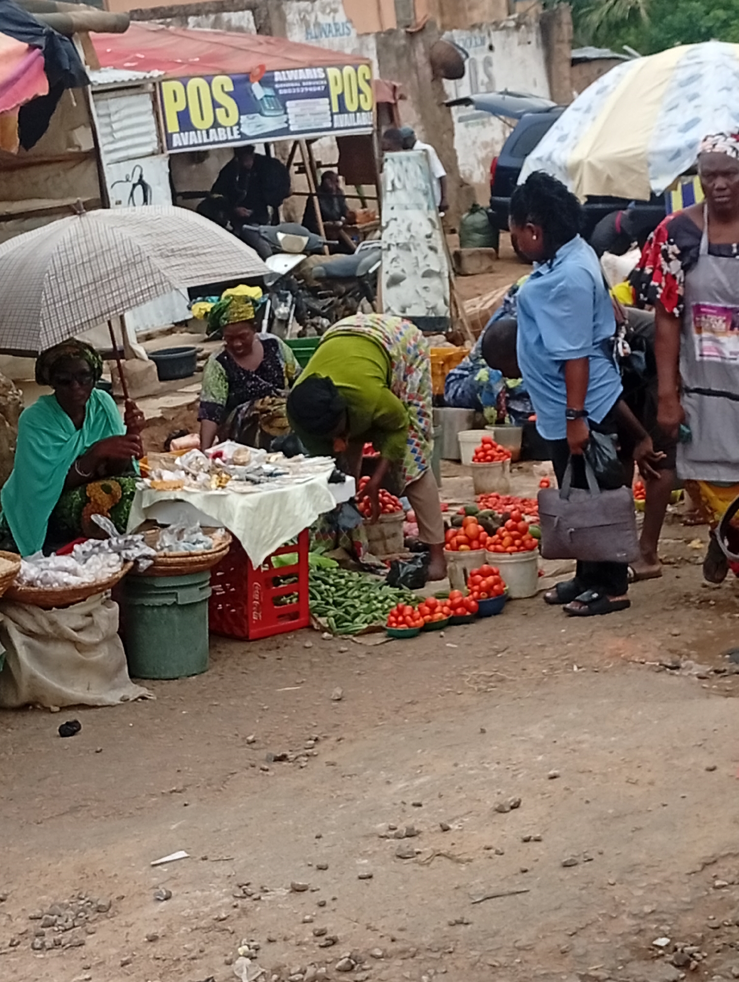 The image depicts a street scene in Maiburiji, Nigeria. Several vendors are selling goods, primarily food items like tomatoes, okra, and dried goods. A woman under a checkered umbrella is selling goods on a table. Another woman in green is stooping to pick items from the ground. A person in a blue shirt is purchasing produce. A red Coca-Cola crate is being used as a display stand. In the background, a sign reads "POS AVAILABLE". People are walking around and interacting. A vehicle is also visible in the background. The ground is dirt.