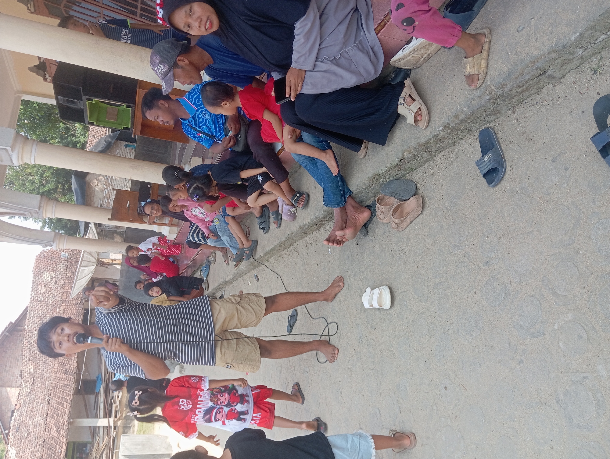 In Dente Teladas, Indonesia, a group of people are gathered outdoors on a concrete surface. A young man in a striped t-shirt and khaki shorts stands and speaks into a microphone, pointing his finger outwards. Several children and adults are seated on a raised concrete step and the ground. Some are looking at the speaker, while others appear to be engaged in their own activities. A large speaker is visible behind the seated individuals, suggesting an event or gathering is taking place. The environment appears to be residential, with a building and vegetation in the background. The overall scene depicts a community gathering with a central speaker engaging the audience.