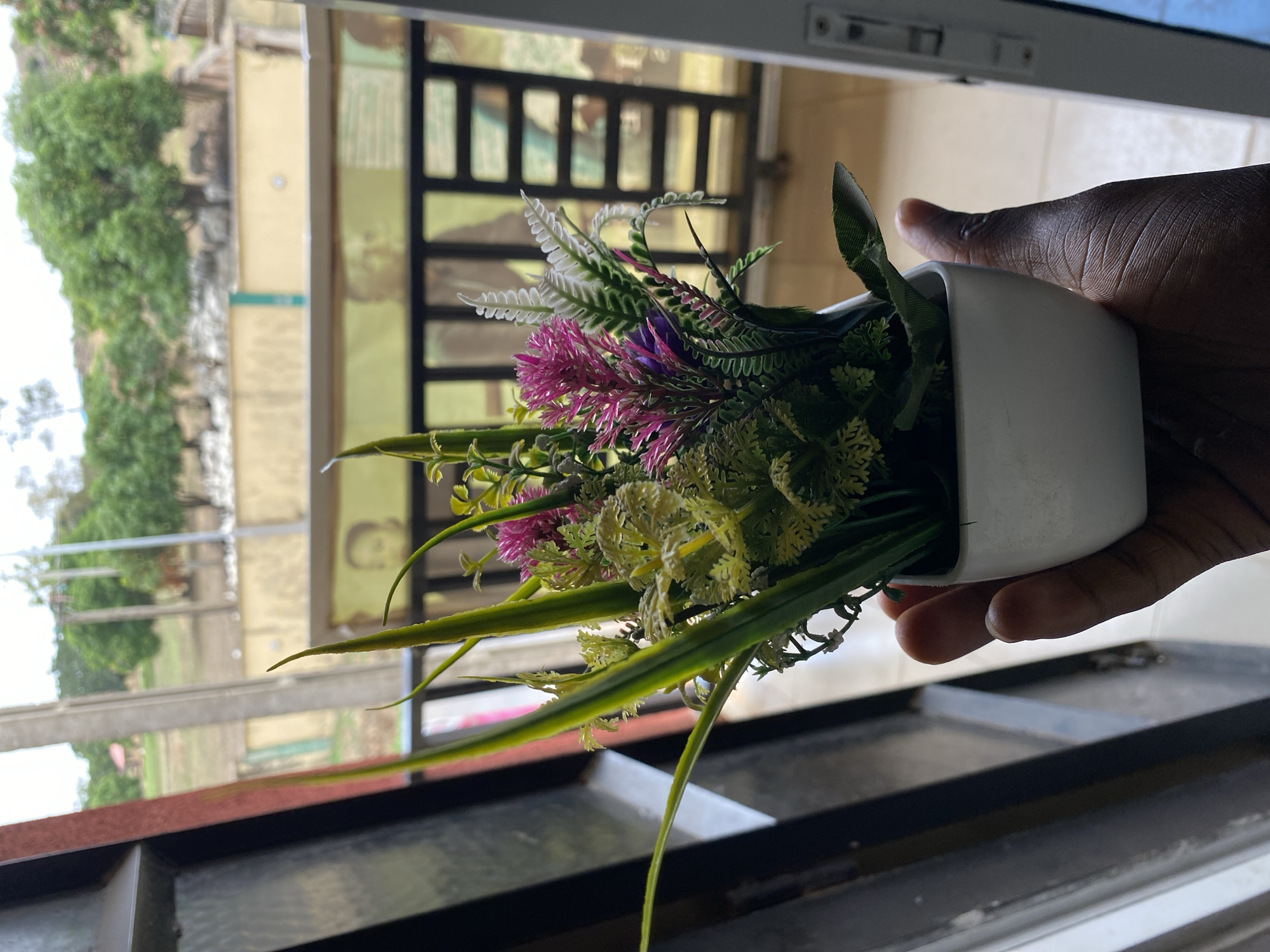 A hand holds a small, white, square pot filled with artificial plants and flowers. The flowers are various colors including purple, white, and yellow. The background shows a window view of greenery and buildings, with a black metal grate partially visible. The location is Bukuru, Nigeria.