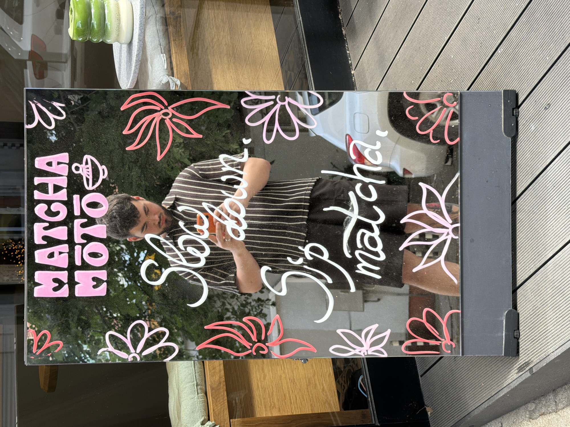 A man wearing a black and white striped shirt and black shorts is standing in front of a reflective board. The board has pink decorative flowers and white lettering. The lettering reads "MATCHA MOTO" at the top and "Slow down. Sip matcha," below. The man is holding a small orange object in his hands and looking down at it. The reflection on the board shows trees, a white car, and a stack of green and white layered drinks on a grey plate. The board is on a wooden deck. The location is in Sofia, Bulgaria.
