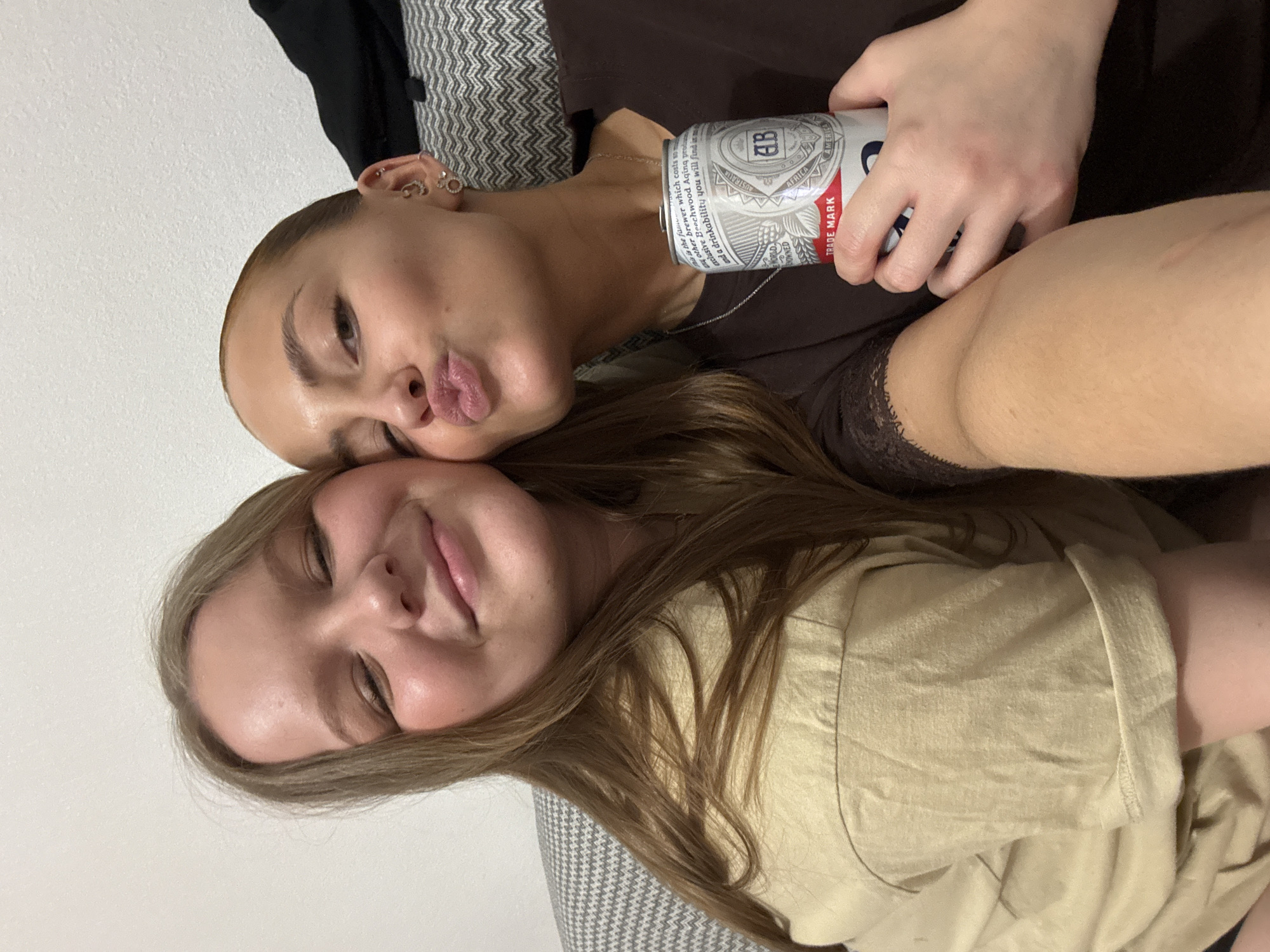 Two young women are lying next to each other on a gray and white patterned couch. The woman on top is holding a can of Budweiser beer. The background is a plain white wall. The exact location in Chaikovsky, Russia, is not discernable from the image.