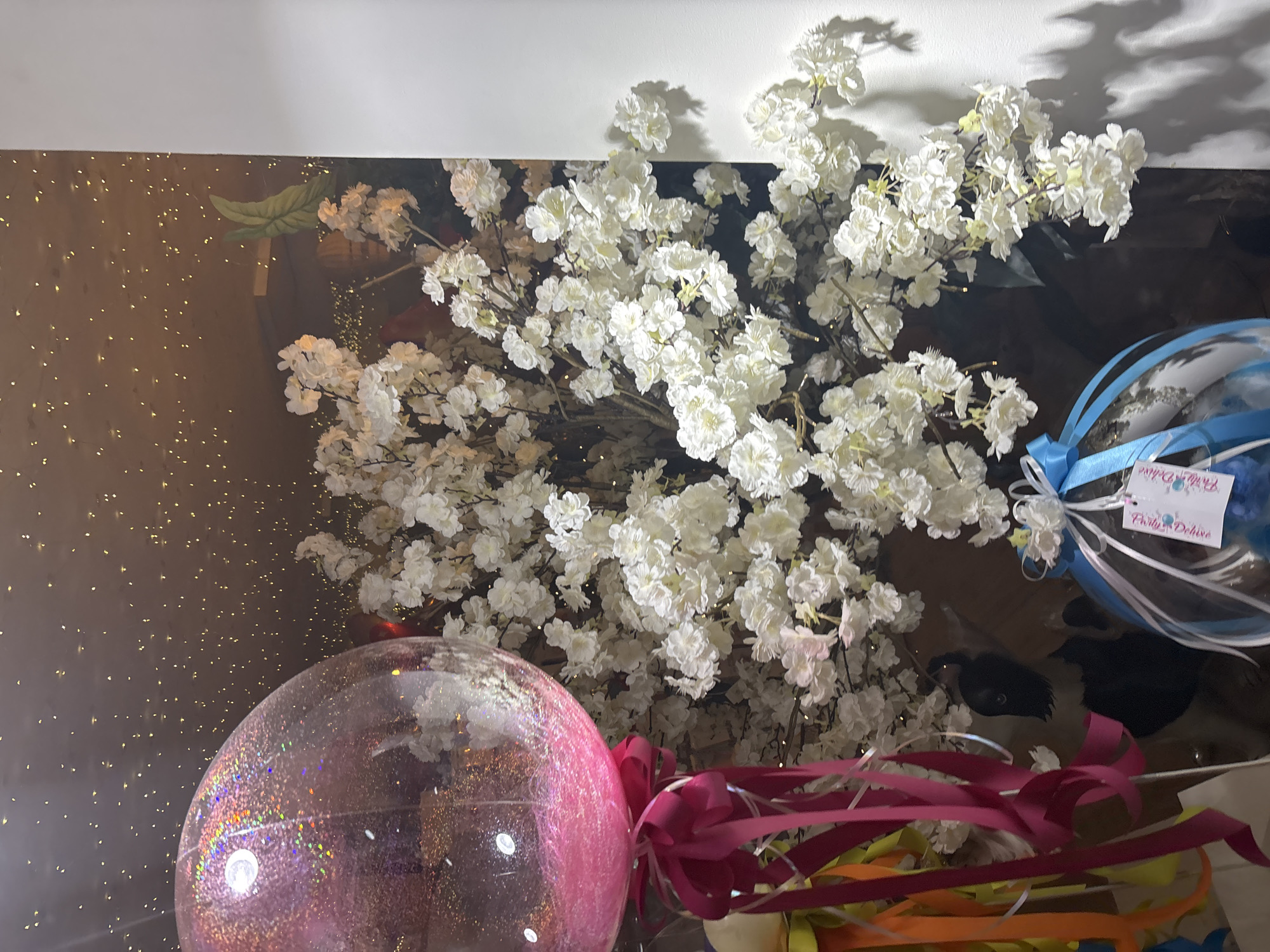 The image shows a display with various decorative items. There is a large bouquet of white flowers, a pink glittery balloon, and several ribbons in pink, orange, and yellow. There is also a blue and white balloon with a tag that reads "Party Deluxe" in a decorative script. The display is positioned against a reflective surface with small, scattered gold-colored lights. The setting suggests a celebration or party arrangement. There are no indications of a specific location in Sofia, Bulgaria.
