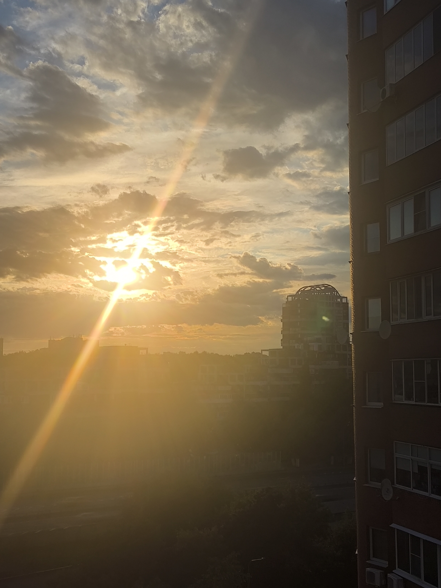The image presents an urban or suburban landscape illuminated by the intense, golden light of a low sun, strongly suggesting either sunrise or sunset. The perspective is elevated, possibly from a building window or balcony.
The sky occupies the upper portion of the frame, filled with a mix of stratocumulus and altocumulus clouds. The sun, a brilliant white-yellow orb, is positioned slightly left of center, partially obscured by clouds, creating a powerful sunburst effect and a prominent, diagonal ray of light (likely a lens flare) that extends towards the lower-left corner, displaying subtle prismatic colors.
In the immediate foreground on the right, a tall, multi-story building with a reddish-brown brick facade is partially visible. It features numerous rectangular windows, some of which appear to be open, and several external air conditioning units are mounted on its side. A small satellite dish is also affixed to the building's exterior.
Further back, in the midground and background, several other buildings are discernible, though largely silhouetted and softened by the strong atmospheric haze and direct sunlight. One notable structure in the midground stands out with a distinctive, domed or lattice-like architectural feature on its roof. The lower central and left parts of the image are heavily saturated with warm, golden light, obscuring finer details but suggesting areas of vegetation and possibly other urban infrastructure under a thick, luminous haze.
The overall color palette is dominated by warm yellows, oranges, and deep gold, especially where the sun's light is most direct, contrasting with the cooler grays and blues of the clouds in less illuminated sections of the sky. There is no visible text in the image, and no specific human activity is depicted, focusing instead on the dramatic natural lighting of the urban environment.