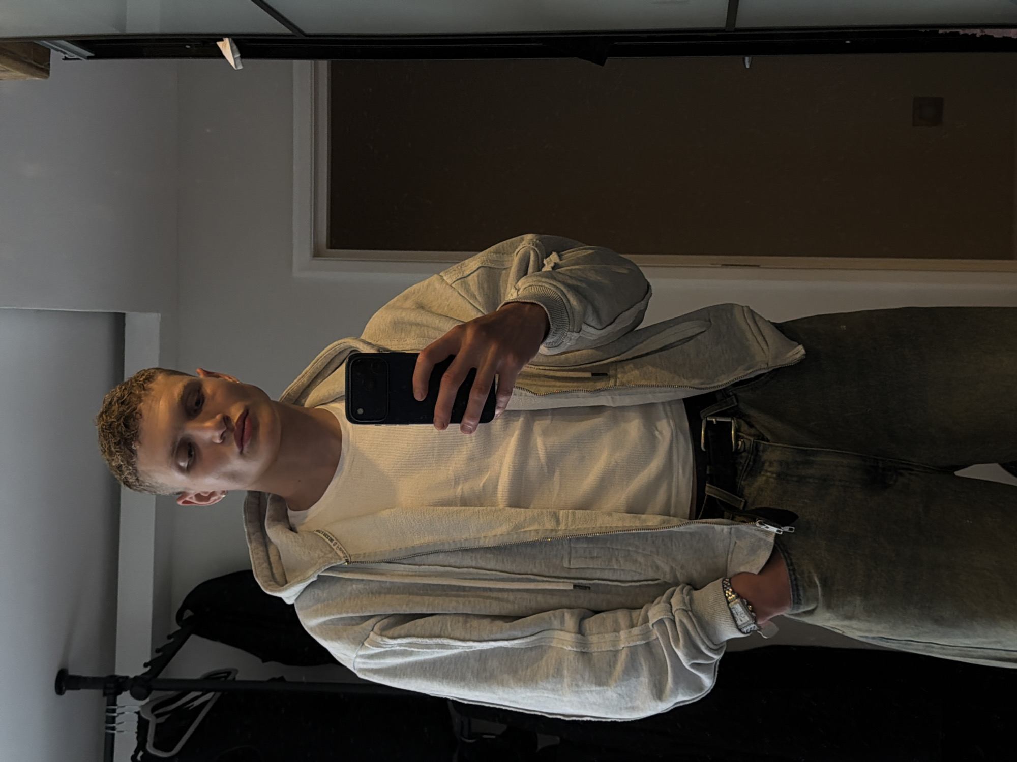 A young man with short, curly blonde hair and light skin is taking a mirror selfie indoors. He is wearing a light grey zip-up hoodie over a white waffle-knit t-shirt, dark green wide-leg jeans, and a black belt. He has a watch on his left wrist and is holding a black smartphone in his right hand. His left hand is in his jeans pocket. The background consists of a closet with clothes hanging on a rack, a doorway, and a full-length mirror. The overall impression is of a casual, domestic setting. The image does not contain any information that would suggest the location is in the city of London, United Kingdom.