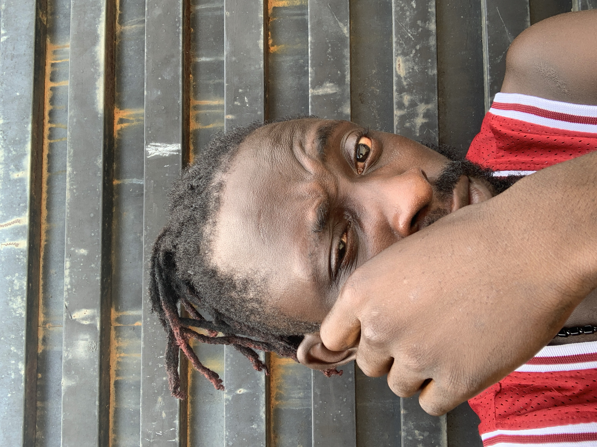 A portrait-oriented image, rotated on its side, features a young man with dark skin, styled dreadlocks, and a beard. He is wearing a red athletic top with white stripes. His right hand is raised, partially covering his mouth and chin area, with his fingers near his ear. He looks directly into the camera with an intense, serious gaze. The background consists of a dark, weathered corrugated metal surface, showing vertical ridges with visible patches of orange-brown rust and other discolorations. The lighting suggests it is daytime, possibly under diffuse light. No text is visible in the image.