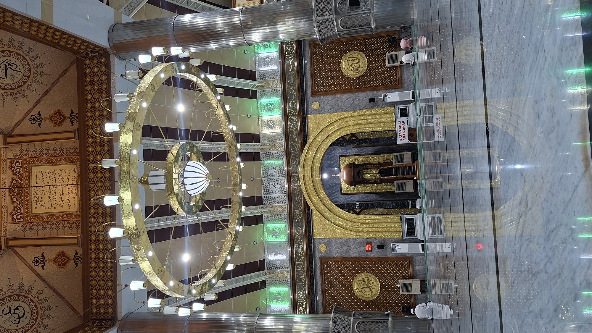 The image shows the interior of a mosque, likely the Grand Mosque of Langsa in Langsa City, Indonesia. The mosque features opulent decoration with intricate Islamic calligraphy on the walls and ceiling. A large, ornate chandelier hangs from the ceiling, illuminating the space. The floor is made of polished marble, reflecting the light and the surrounding architecture. There are several prayer mats on the floor, and a few individuals are seen praying. The mihrab, a niche in the wall indicating the direction of Mecca, is prominently featured in the center, adorned with golden arches. Air conditioning units are visible on the floor, suggesting a modern facility. A sign in red and white text is partially visible, likely providing instructions or information. The overall impression is one of grandeur and religious solemnity.