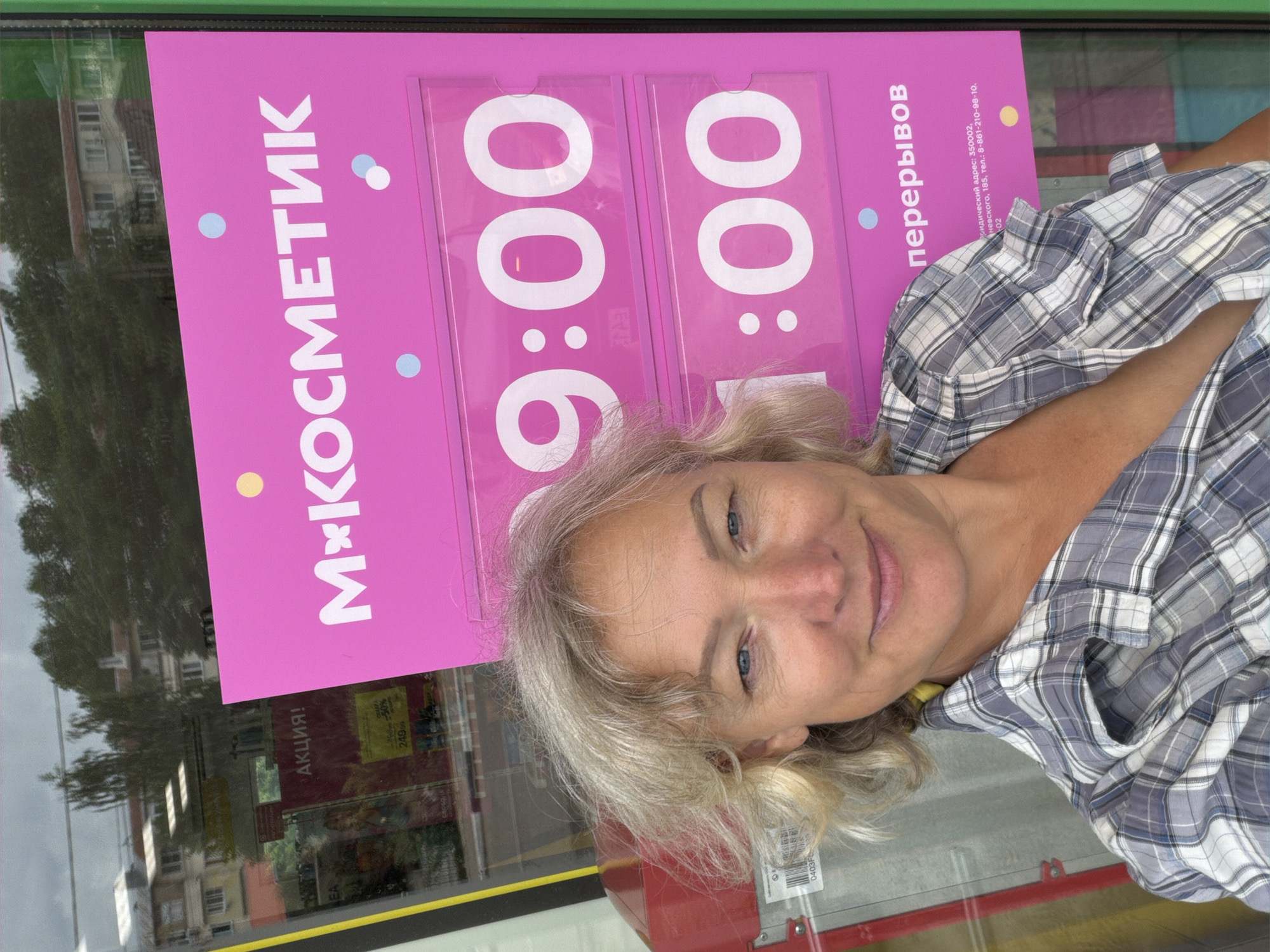 A woman with blonde hair and blue eyes smiles at the camera, standing in front of a pink sign with white text. The sign has the text "МХ КОСМЕТИК" and below it, two time slots: "9:00" and "1:00". Further down, it says "перерывов". The sign also displays an address and phone number. The woman is wearing a grey and white checkered shirt. Reflections in the window behind her show buildings and trees, suggesting an outdoor urban environment. The image is taken outdoors, possibly in front of a shop. The location is identified as Shepsi, Russia.