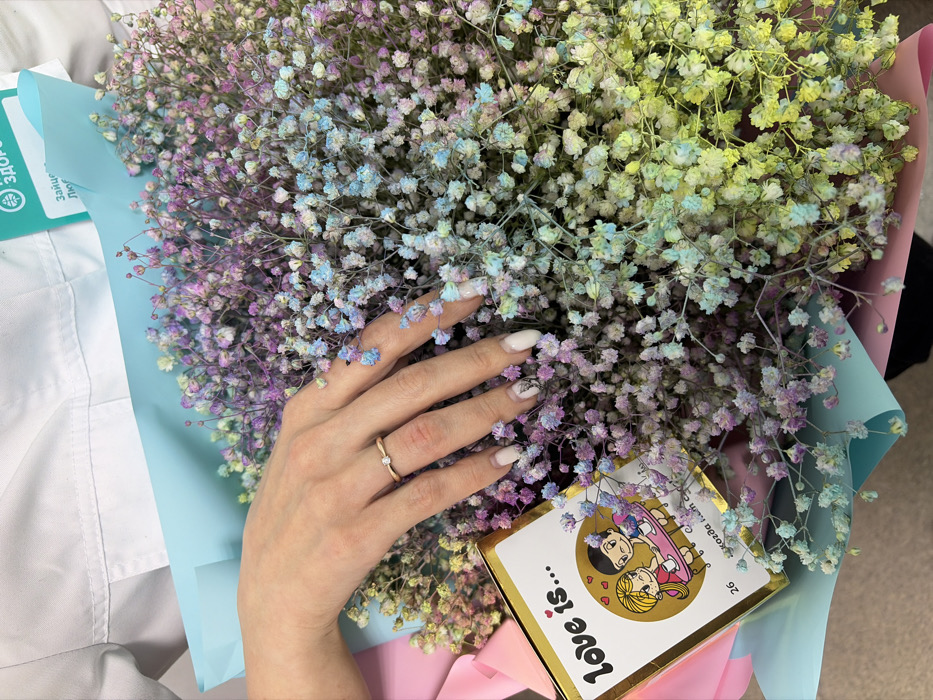 A close-up, top-down view captures a person holding a large, dyed gypsophila flower bouquet and a small gift. The flowers exhibit a multi-color gradient, transitioning from light purple to light blue and yellow-green, and are wrapped in light blue and pink paper or film. A female left hand, with manicured nails featuring white polish and black abstract designs on some fingers, rests on the bouquet. A gold-toned ring with a single clear stone is visible on the ring finger. Nestled within the flowers is a rectangular "Love is..." branded box with a gold border, displaying the text "Love is..." and the number "26." A portion of a green label with Russian text, including "Здоровье" (Health) and "Люди" (People), is visible in the upper right corner, against what appears to be a white uniform, suggesting a medical or pharmaceutical setting. The scene is located in Chaikovsky, Russia.