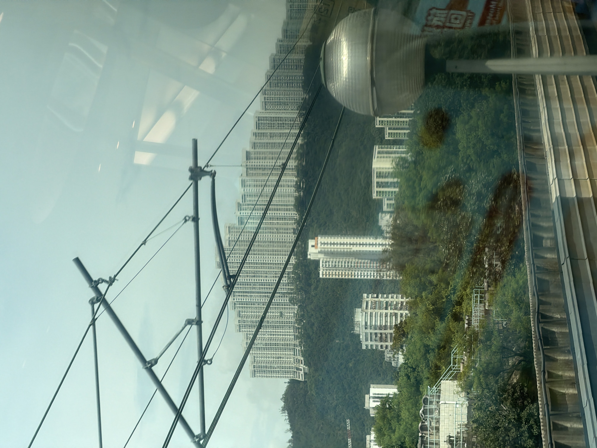 The image shows a panoramic view of residential buildings on a hillside in Hong Kong. The buildings are tall and densely packed, characteristic of urban Hong Kong. The scene is viewed through a window, with overhead cables and a reflection of a light fixture visible in the foreground. The hillside is covered with lush green trees, contrasting with the white and gray facades of the apartment blocks. The sky is overcast. This view is likely from a public transport system like the MTR, given the presence of overhead electrical cables.