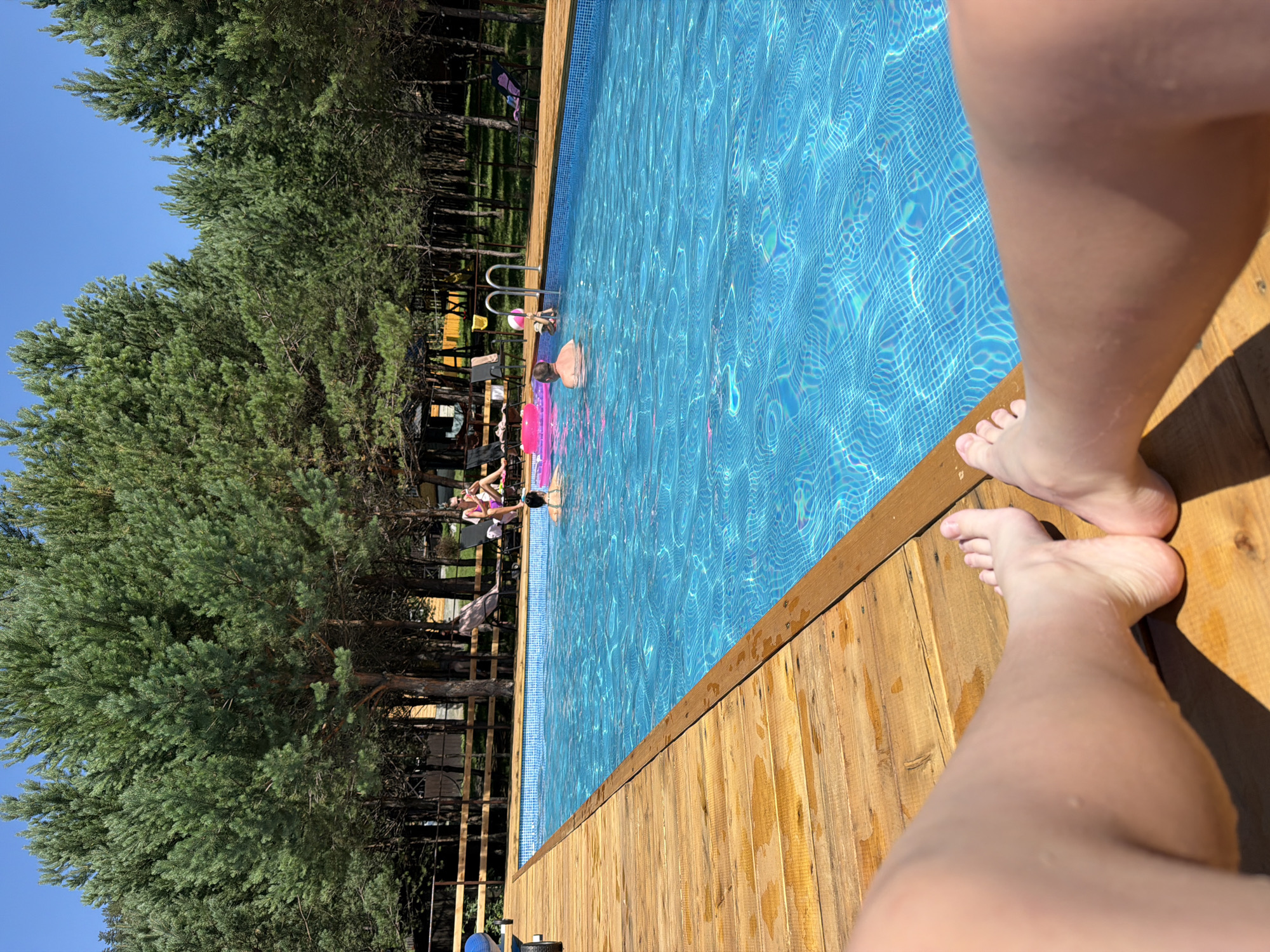 The image depicts a swimming pool area in Elekmonar, Russia, with a focus on a wooden deck in the foreground. Several people are visible in the pool, including adults and children. Some are swimming, while others are relaxing on inflatable toys. A pink inflatable ring is floating in the water. In the background, there is a dense forest of pine trees. The sky is clear and blue. The wooden deck appears to be constructed from planks and is wet in places, suggesting recent use. The overall scene conveys a sense of a recreational summer day.