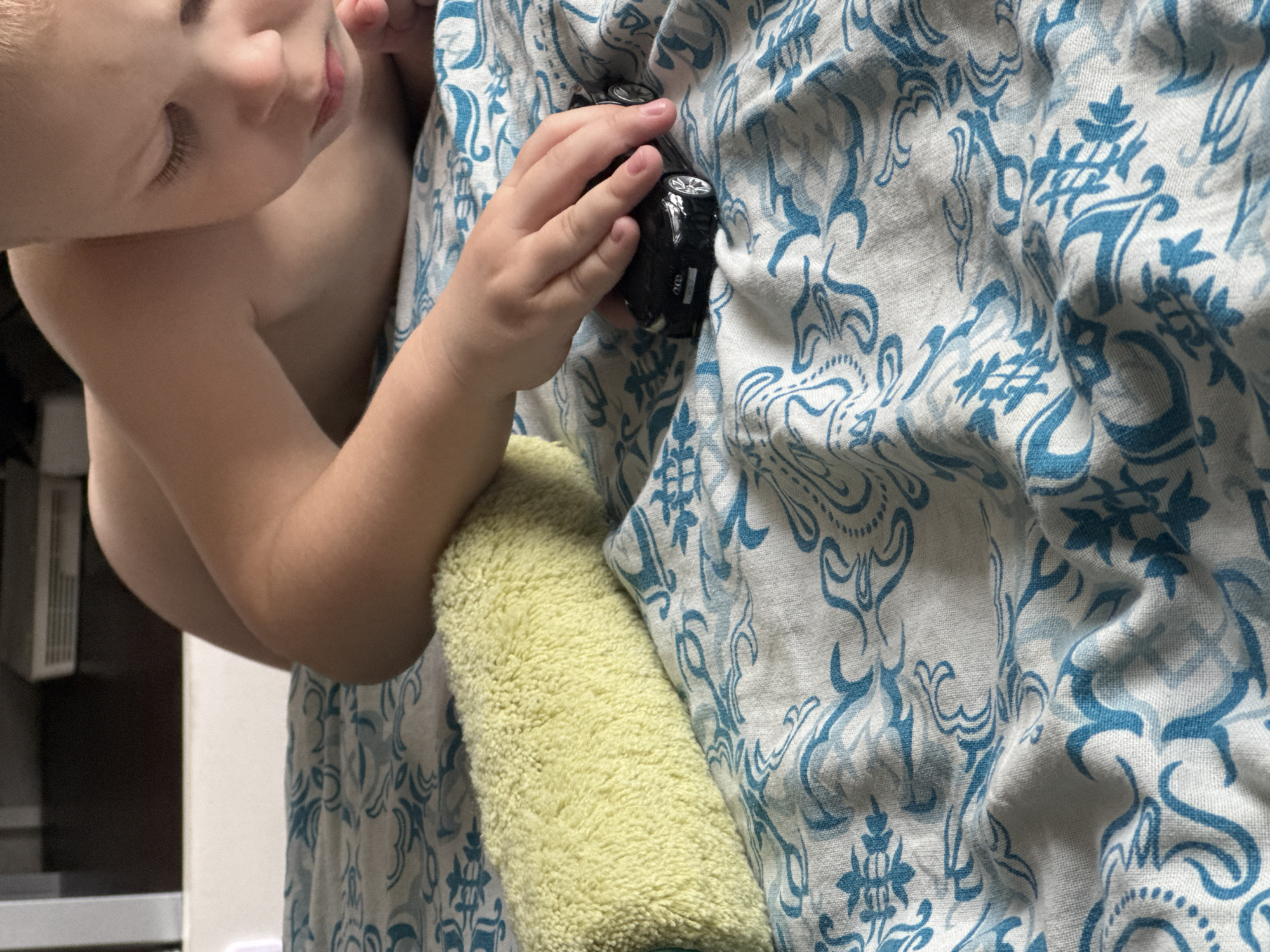 A close-up shot captures a young child's head and arm as they interact with a miniature black car. The child, appearing to be a toddler, is seen from the side, with their face partially obscured and their eyes closed or looking down. Their delicate eyelashes are visible. The child's bare shoulder is in frame, and their arm is extended, holding the toy car. The car is black with silver accents on the wheels and what appears to be a logo or emblem on its side.
The child is nestled against a textured surface that resembles a soft, pale yellow towel or blanket. Behind the child and the towel is fabric with a repeating pattern of blue, stylized floral or paisley designs on a white background. The fabric is draped and creased, suggesting it might be a bedspread or upholstery.
The overall mood is intimate and tranquil, possibly indicating a moment of play or rest. The lighting appears to be indoor, with no strong indicators of the time of day or specific weather conditions. The background is blurred, but hints of what might be furniture or appliances in a domestic setting are discernible on the left side of the image. There is no visible text in the image. The scene evokes a sense of childhood innocence and gentle interaction with toys.