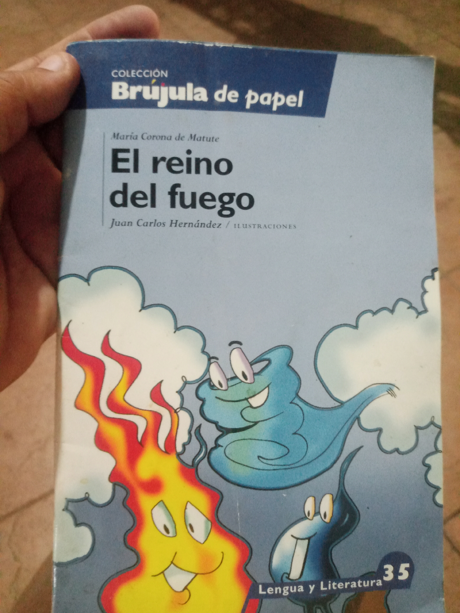 A person's hand is holding a light blue book with a dark blue band across the top. The book is titled "Coleccion Brujula de Papel" and "El reino del fuego" by Maria Corona de Matute. The book cover features cartoon illustrations of fire and blue creatures with wide eyes and open mouths. The number "35" is visible in the bottom right corner, along with the words "Lengua y Literatura". The background is out of focus and appears to be a wooden surface. The image is taken at an angle, with the top of the book tilted away from the viewer. There are no discernible landmarks or indications of the city of Santa Ana, Venezuela.