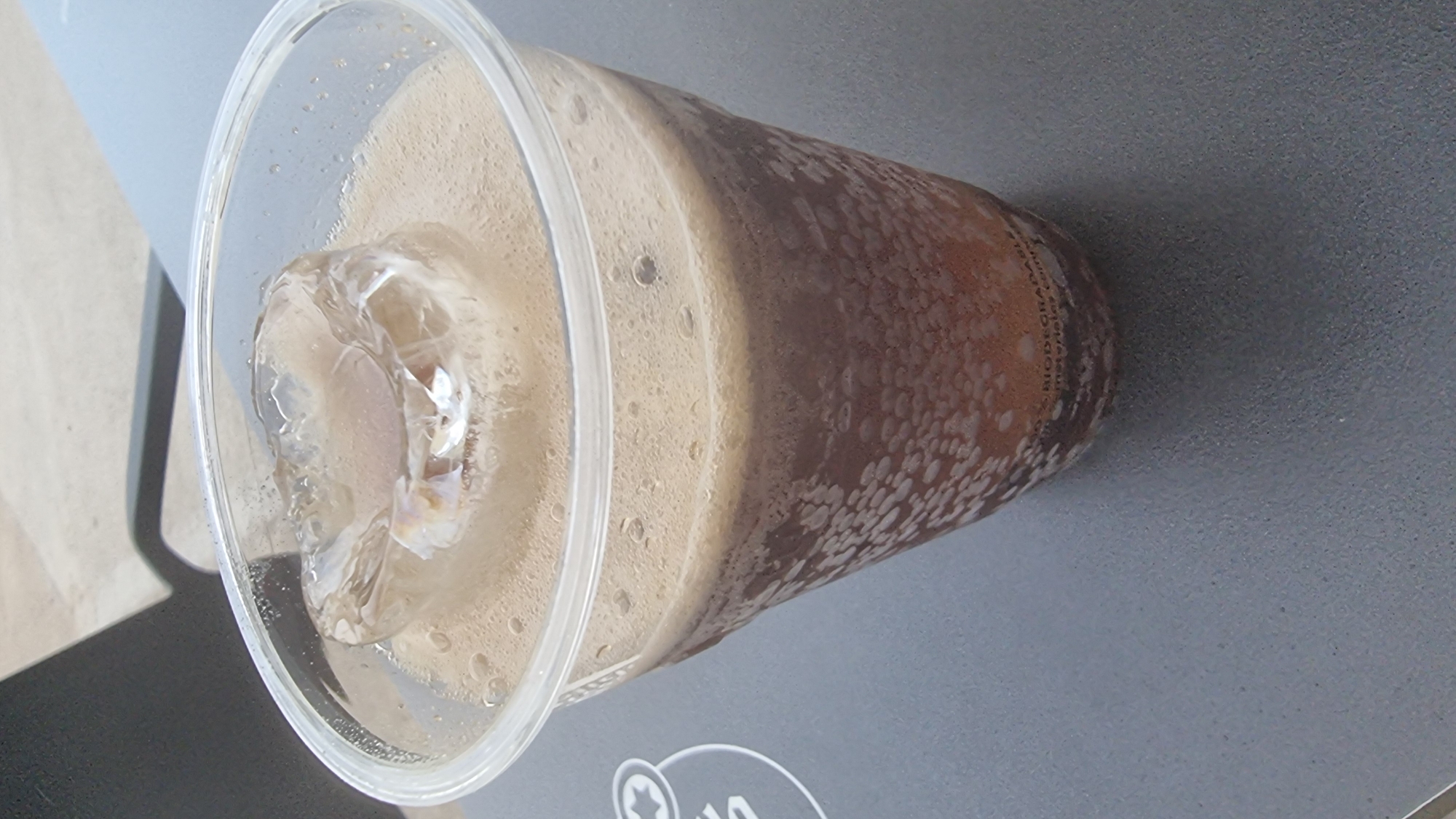 A close-up shot shows a clear plastic cup filled with a dark, fizzy beverage and ice. The drink is topped with a foamy layer and a large piece of ice that appears to be melting. The cup is positioned on a textured gray surface, possibly a table. Some text, partially obscured by the beverage and condensation, is visible near the bottom of the cup, appearing to say "BIODEGRADABLE" and "materials.com". The background is out of focus, with hints of a shadow and an outdoor setting. There is no visible indication of the city of Olot, Spain.