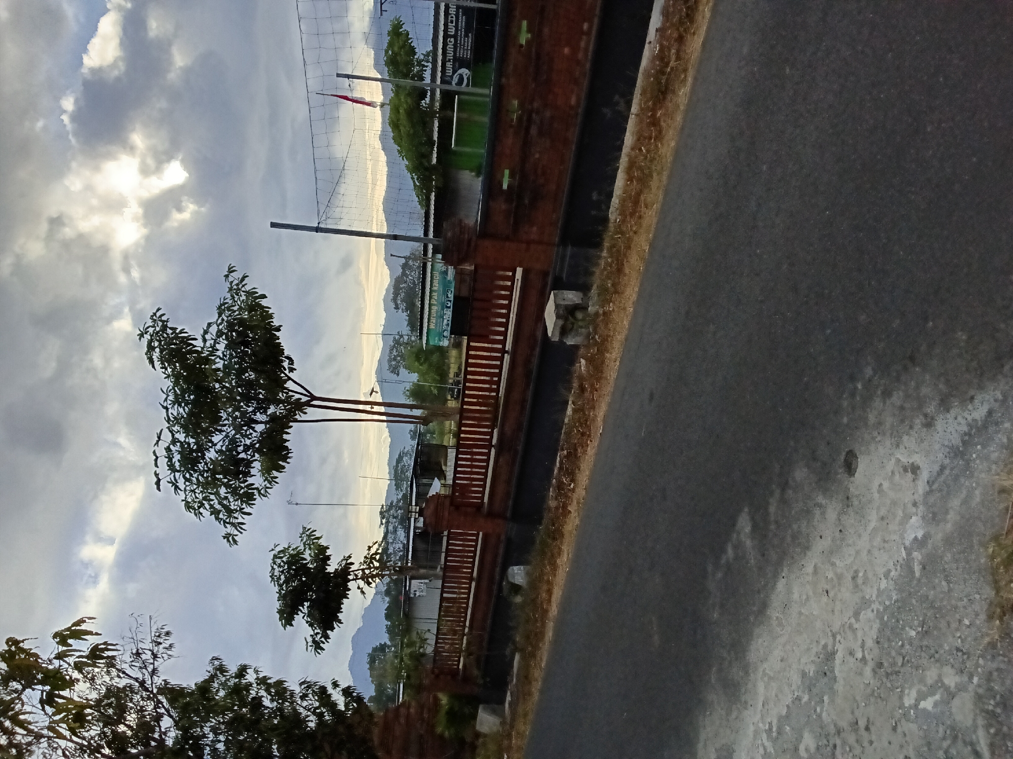 The image depicts a scene in Bendorejo, Indonesia, likely a rural or semi-urban area. In the foreground, an asphalt road runs diagonally from the bottom left to the bottom right. The road has some patches of lighter colored material on its surface. To the right of the road, a low brick wall with a wooden fence on top runs parallel to it. Beyond the wall, a grassy area is visible, and further back, there are buildings with metal roofs. A prominent sign on one of the buildings reads "WARUNG PAK KANCIL" which translates to "Pak Kancil's Stall". Another sign next to it has text that is partially obscured. A volleyball net stretches across the background, suggesting a recreational area. In the far distance, a range of mountains can be seen under a cloudy sky. The clouds are heavy and dramatic, with some sunlight breaking through in the upper center of the frame. Several trees are scattered throughout the scene, with one tall, slender tree standing out near the volleyball net. A red and white flag, likely the Indonesian flag, is visible on a pole behind the volleyball net. The overall lighting suggests it might be late afternoon or early morning. There are no people visible in the image.