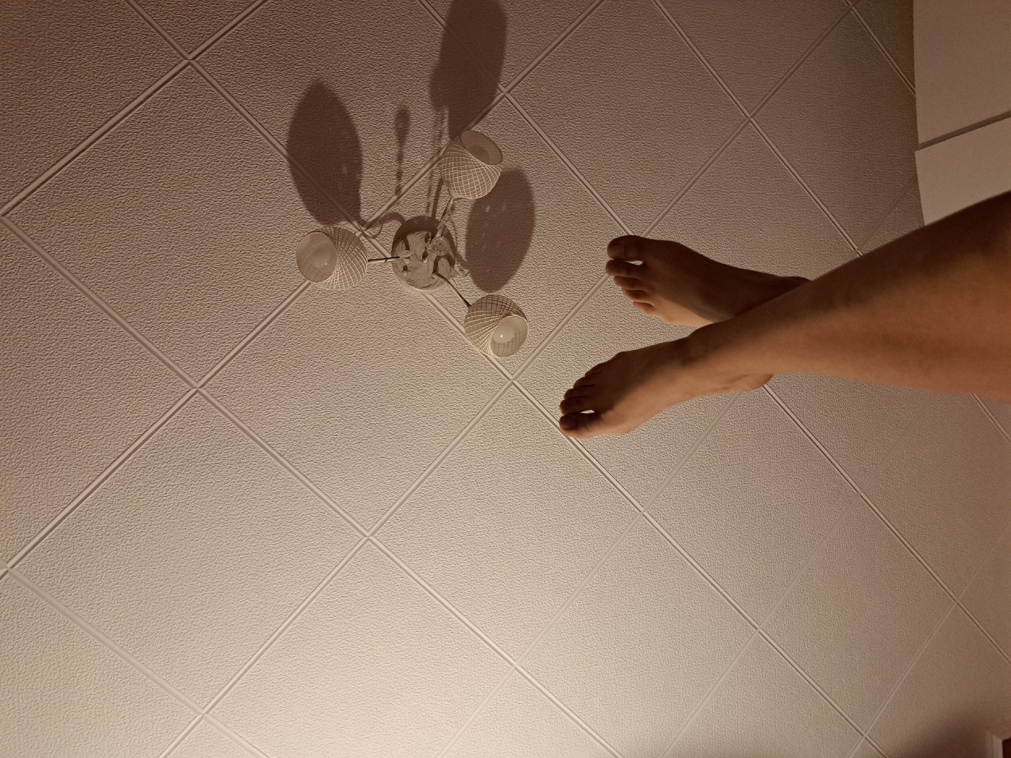 A low-angle shot captures a white textured ceiling with a diamond-shaped pattern. A three-bulb light fixture with white basket-like shades hangs from the ceiling, casting shadows. In the foreground, two bare human feet are crossed. The left leg extends upward from the bottom left of the frame, and the right leg crosses over the left, pointing towards the top right. The scene appears to be indoors, possibly a bedroom or living room.
