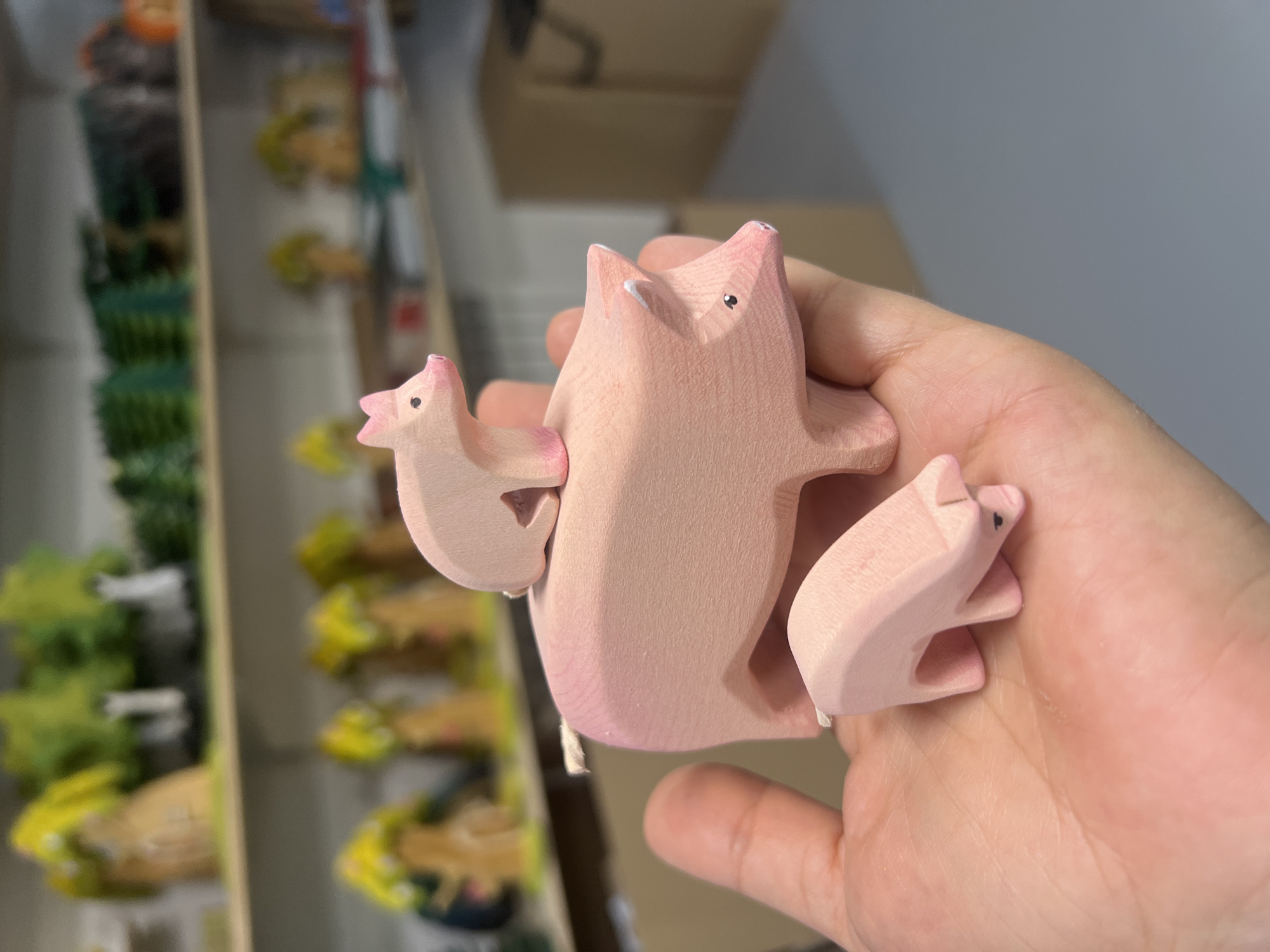 Three pink, wooden pig figurines are held in a person's left palm and hand. The largest pig is lying down on its side, facing right, with its head slightly raised. A smaller pig is resting on the back of the larger pig, facing left. An even smaller pig is standing in the palm of the hand, facing right and with its head down. The background is blurred and shows shelves with various blurred objects, possibly toys or craft supplies, and a cardboard box. The image appears to be taken indoors in a retail or workshop setting.