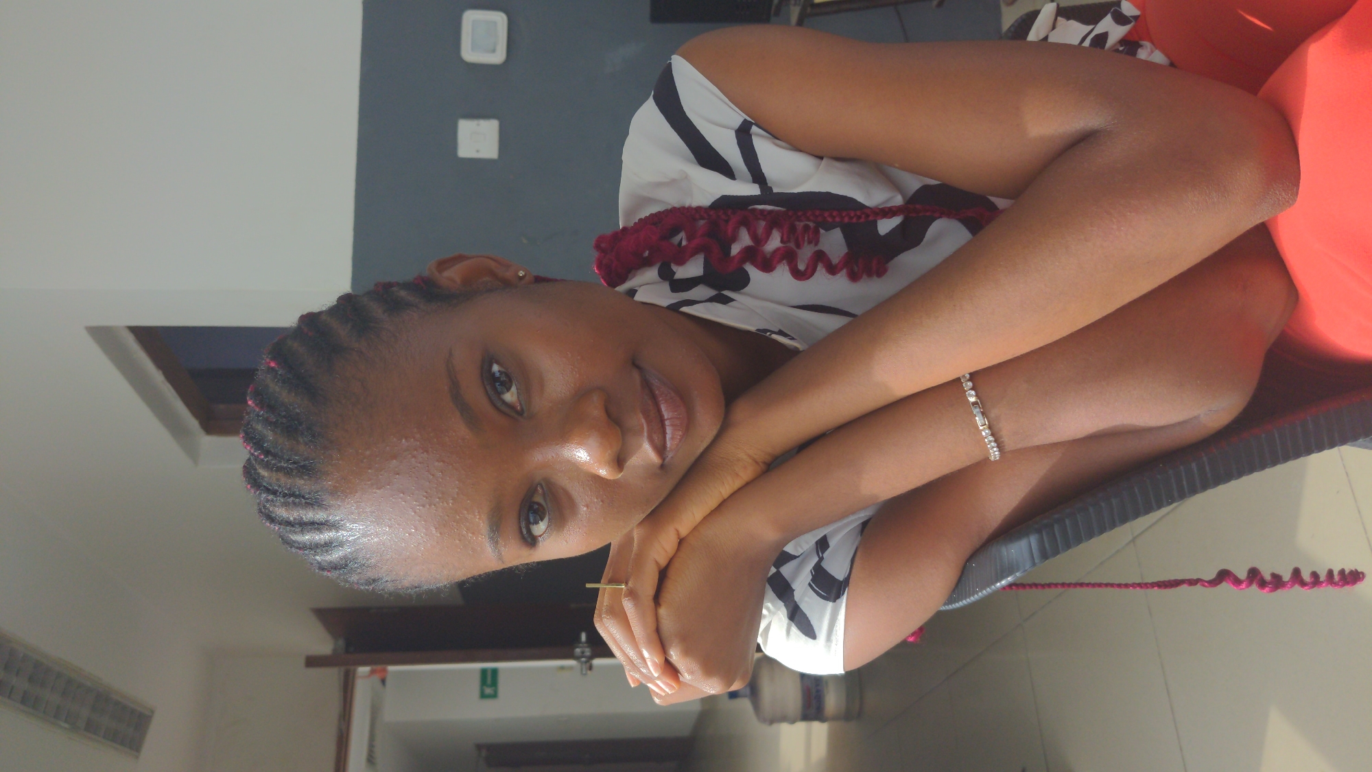 A young Black woman is resting her head on her arms, which are folded on a surface. She is wearing a light-colored sleeveless top with dark geometric patterns. Her hair is braided with some red highlights woven in. She has a bracelet on her left wrist and a ring on one of her fingers.
The background is blurred, but it appears to be an interior space. There is a gray and white wall with a couple of light switches. There is a glimpse of a wooden door and a water dispenser.
Without explicit landmarks, pinpointing the location in Abuja, Nigeria, is not possible, but the interior setting suggests a domestic or office environment within the city.