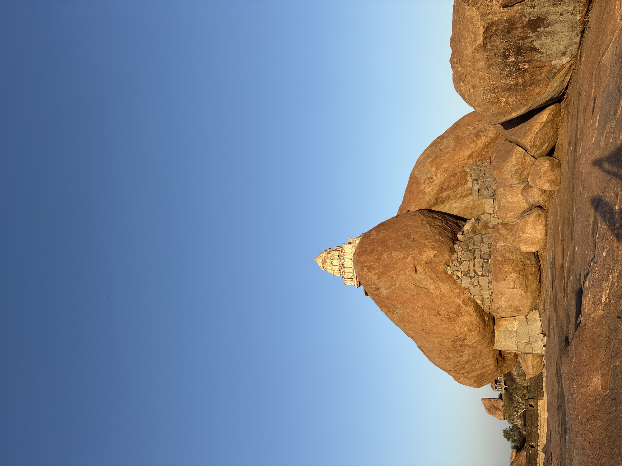 The image shows a view of the Hanuman Temple at Hampi, located in Kamalapura, India. The temple is built on top of a large rock formation. The sky is a clear blue with a slight gradient. The rock formations are primarily reddish-brown. In some parts, one can see evidence of structures made from stones that might be linked to the temple. The sun is on the rocks, creating shadows on the rock surfaces.