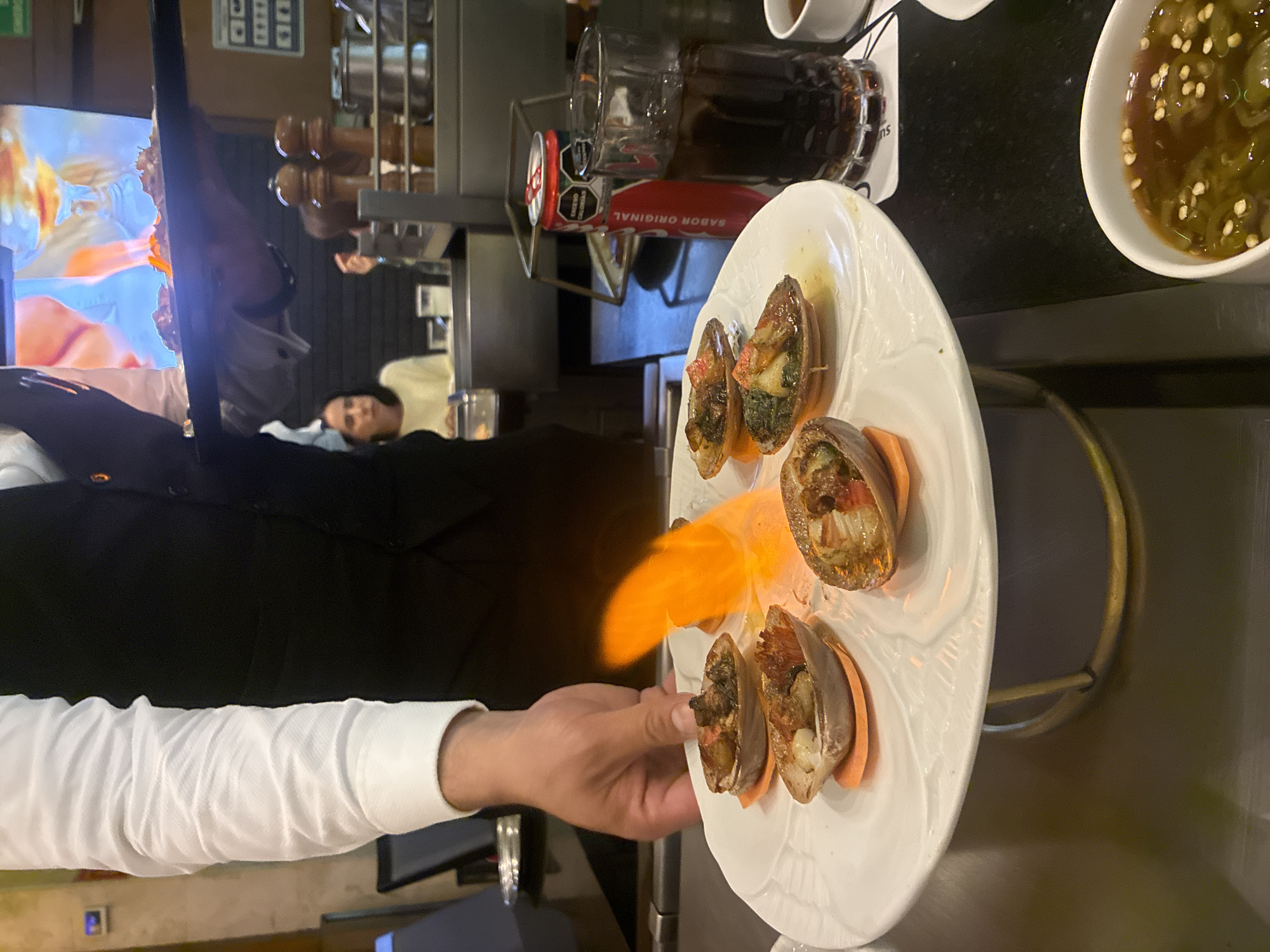 A server in a white shirt and black vest is presenting a plate of abalone shells that are being flambéed with a bright orange flame. The plate, raised on a gold stand, contains several abalone shells filled with food, some of which are visibly charred from the flame. In the background, a woman sits at a table. To the right of the plate, a can of Coca-Cola and a glass of dark liquid are visible, along with a small white bowl containing a sauce with chopped green peppers. The setting appears to be a restaurant, likely in Jesús del Monte, Mexico, given the context provided. The lighting is dim, with the bright flame being the most illuminated element on the food.