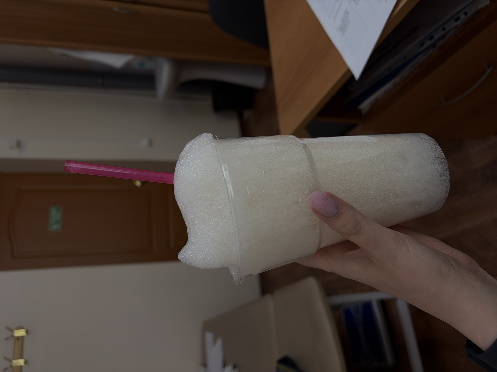 A person holds a clear plastic cup filled with white, foamy liquid and a pink straw in their right hand. The person's hand is fair-skinned with a pink fingernail. The background includes a light brown door, beige wall, and office furniture. The cup is overflowing with the foamy liquid. The location is suggested to be Chaikovsky, Russia.
