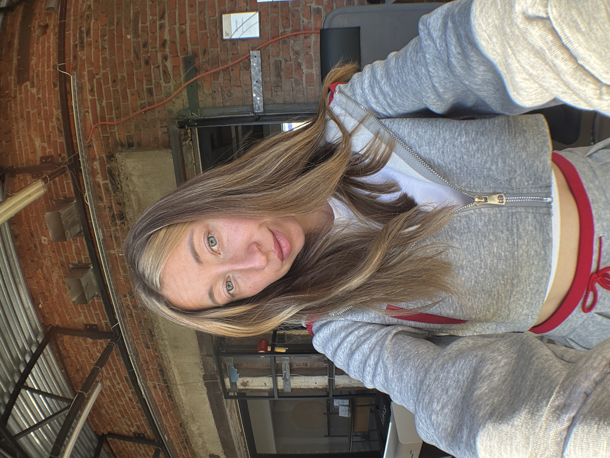 A young woman with long, blonde-streaked brown hair and green eyes is taking a selfie outdoors. She is wearing a gray cropped zip-up hoodie over a white top, and a gray sweatpant with red trim. Her hands are visible in the foreground, suggesting she is holding a phone. The background shows a brick wall with metal roofing and industrial elements, possibly indicating an outdoor commercial or industrial space. The lighting suggests it is daytime. There is no indication that this image was taken in Chaikovsky, Russia.
