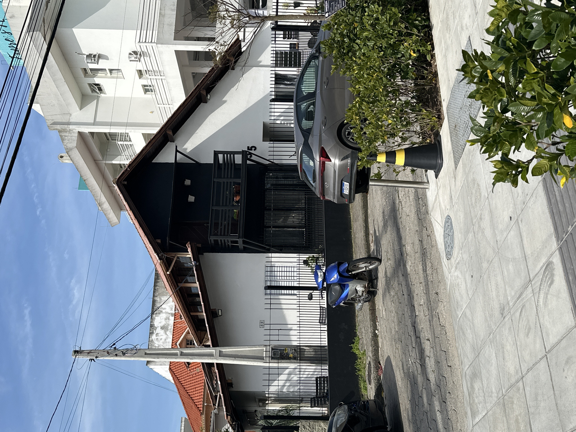 The image depicts a street scene in Florianópolis, Brazil, under a clear blue sky with scattered clouds. In the foreground, a grey hatchback car is parked facing away from the camera, its rear to the right. Beside the car, a dense green bush partially obscures a black traffic cone with yellow stripes and a metal utility cover on the sidewalk.

Behind the car and bush is a street paved with cobblestones, on which a blue motorcycle is parked. A black metal fence runs along the property line. Behind the fence, a two-story house with a white facade and a dark-colored upper section is visible, featuring a sloping red-tiled roof. A balcony with black railings is present on the upper floor. The number "35" is displayed on the front of the house.

Further in the background, a multi-story white building with balconies and windows stands tall. Power lines crisscross the sky, suspended from a concrete utility pole positioned to the left of the house. The environment appears to be a residential or commercial area within the city. There are no people visible in the image.