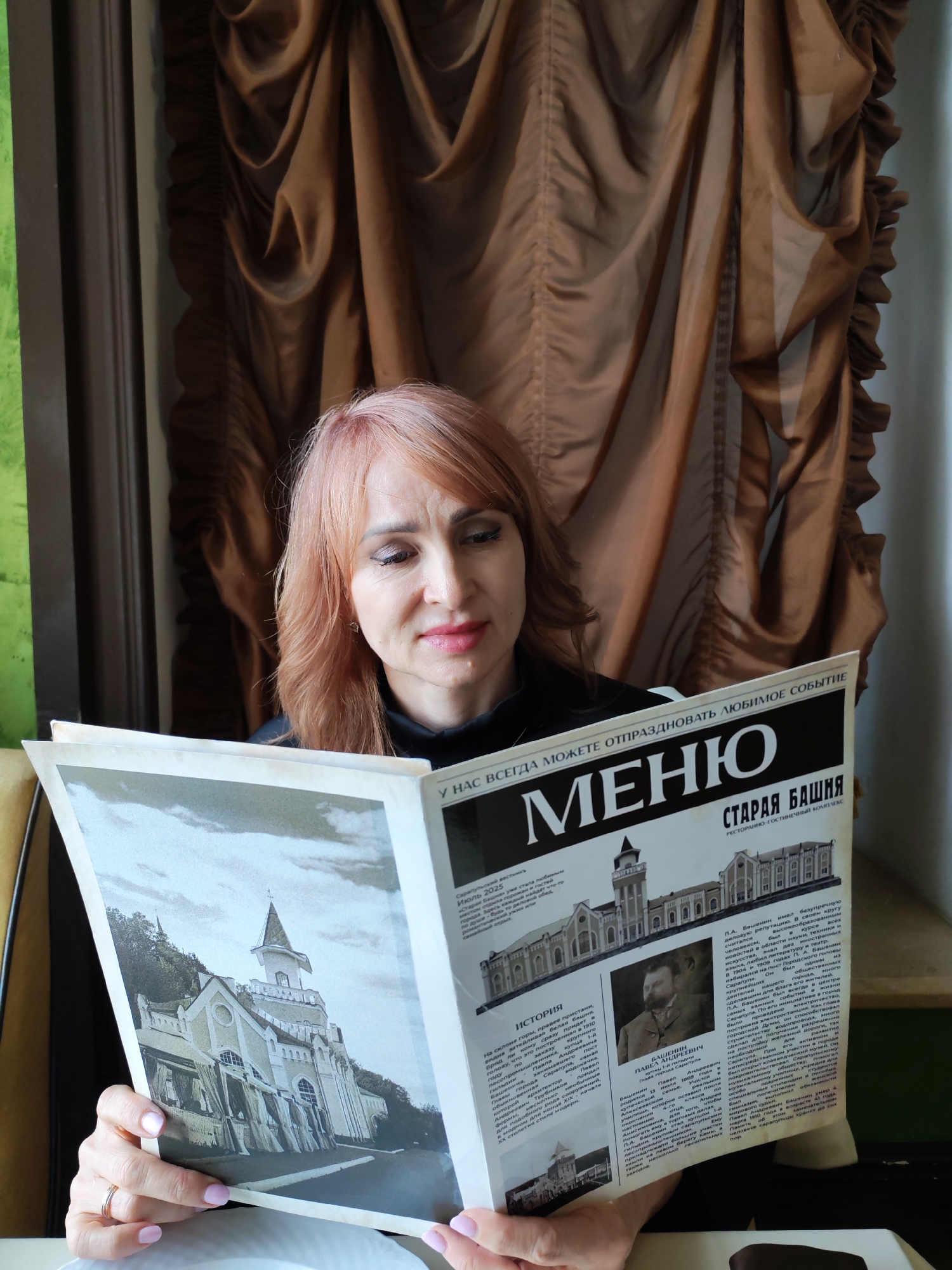 A woman with light pink hair is sitting indoors, reading a newspaper or magazine with the headline "МЕНЮ" (MENU) and "СТАРАЯ БАШНЯ" (OLD TOWER) printed on it. The newspaper features an image of a building, likely the "Old Tower" of Sarapul, Russia. The newspaper also contains text in Russian. The woman is holding the newspaper with both hands. She is wearing a black top. Behind her are brown drapes. A window with green glass is visible to the left. The woman is likely in a restaurant or café.
