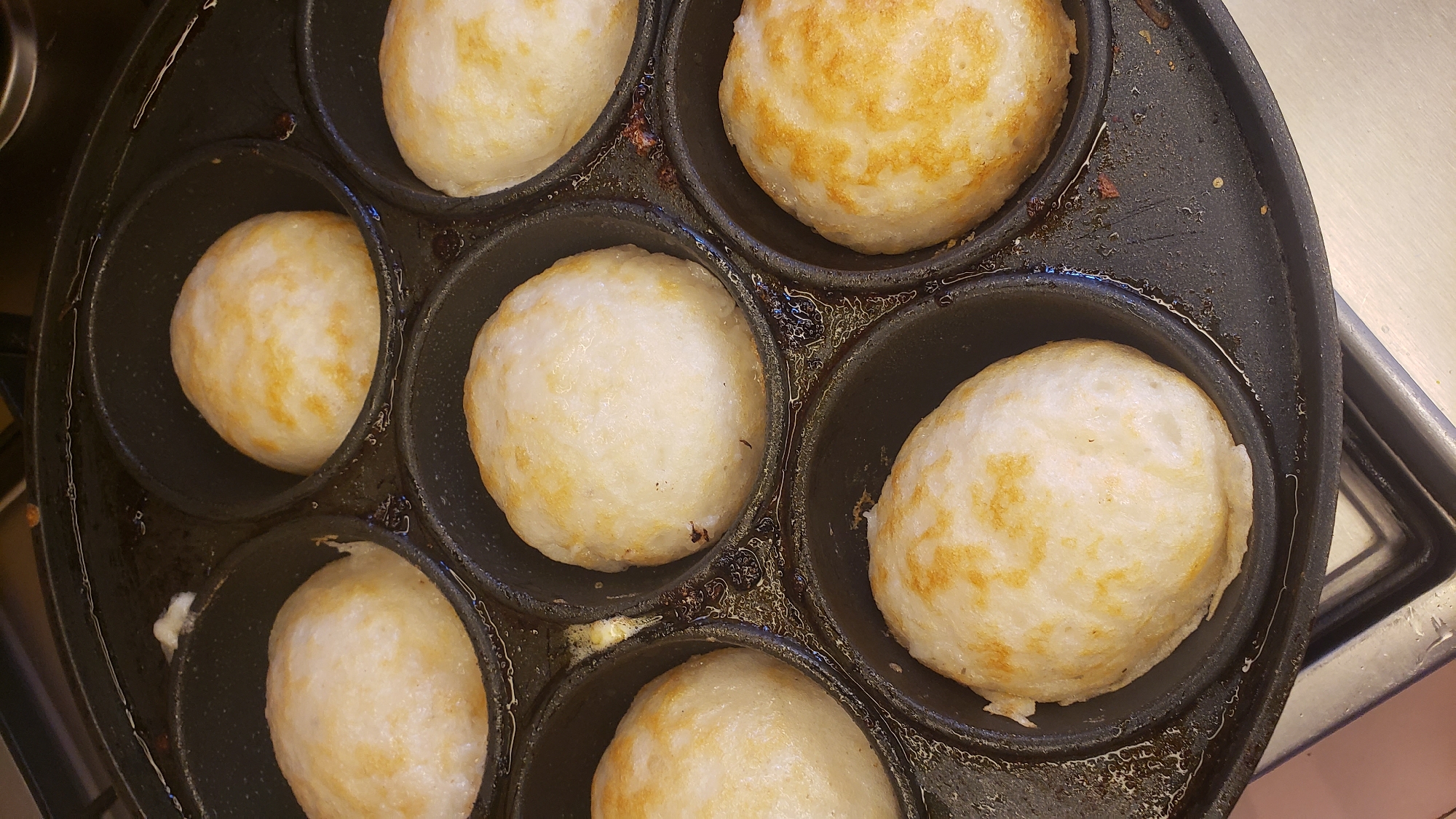 A top-down, close-up, eye-level shot shows a round, black, non-stick paniyaram pan with seven spherical, pale yellow, cooked batters filling each of its indentations. The paniyaram, a South Indian dish, is commonly cooked in this type of pan. The surface of the pan has a slight sheen from oil or butter, with some dark brown residue visible in the crevices between the indentations. The batter has a slightly uneven, porous texture with some areas showing a light golden brown coloration, indicating they are well-cooked. The pan appears to be placed on a stainless steel stovetop, which is partially visible in the lower portion of the frame. The background is blurred, suggesting a focus on the food item. The scene is entirely devoid of people and takes place indoors, likely in a kitchen.