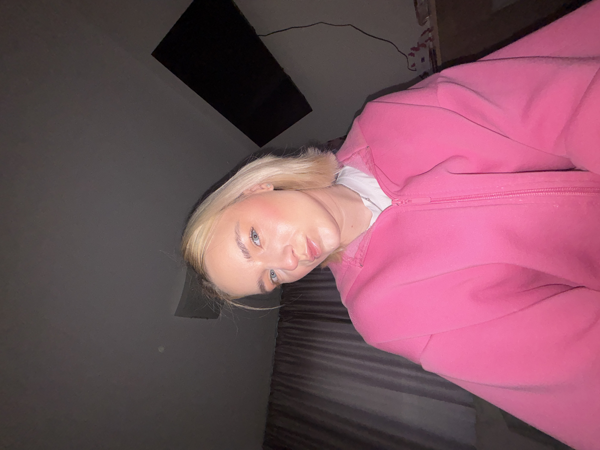 A woman with blonde hair and blue eyes is wearing a pink hooded zip-up sweatshirt. She is lying on a dark-colored bed. The setting appears to be indoors, with a dark gray wall in the background and a black rectangular object mounted on the wall. There is no evidence to suggest that this image was taken in Vsevolozhsk, Russia.